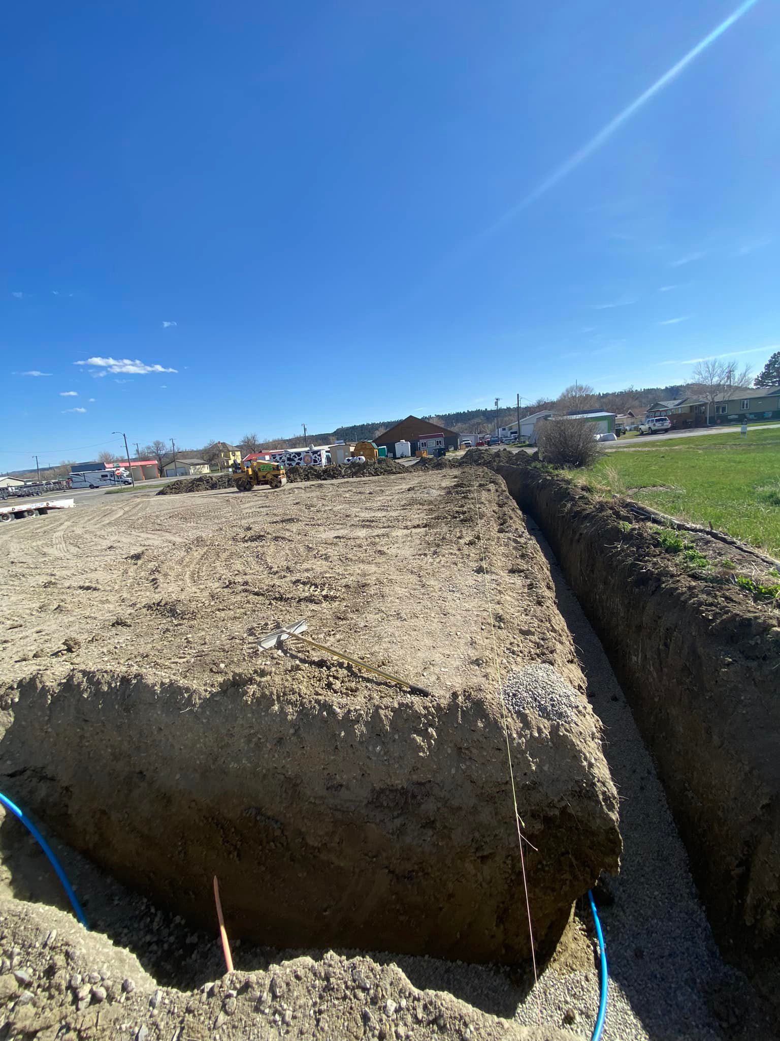 Construction site with a long trench dug in dirt under a blue sky, gravel base, and blue pipe visible.