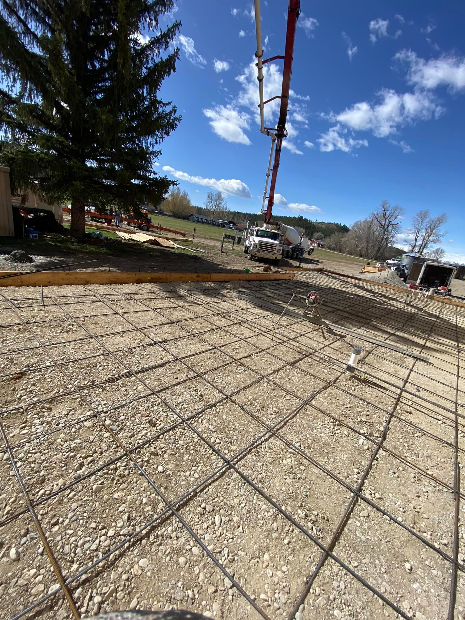 Construction site with concrete pump truck pouring concrete over rebar grid on a gravel base under a blue sky.