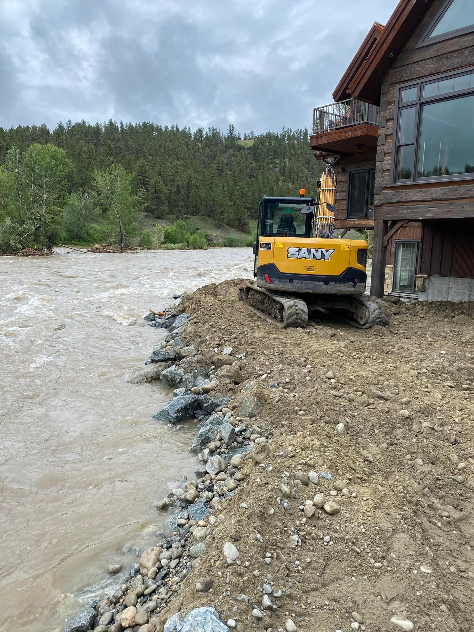 Excavator on a riverbank next to a building, the river is overflowing, cloudy sky.