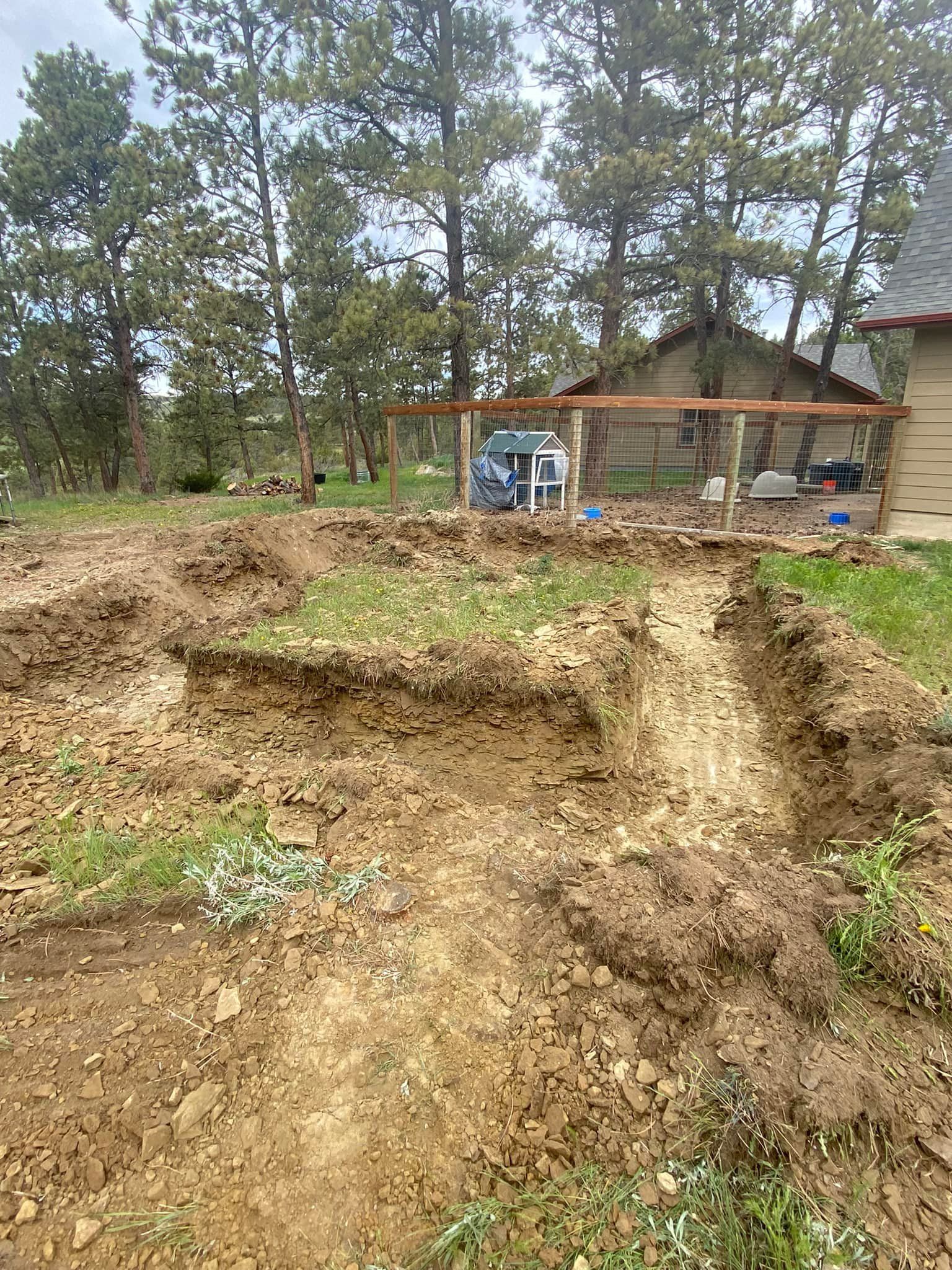Dirt trenches in a yard, trees in the background, a small structure and a car.