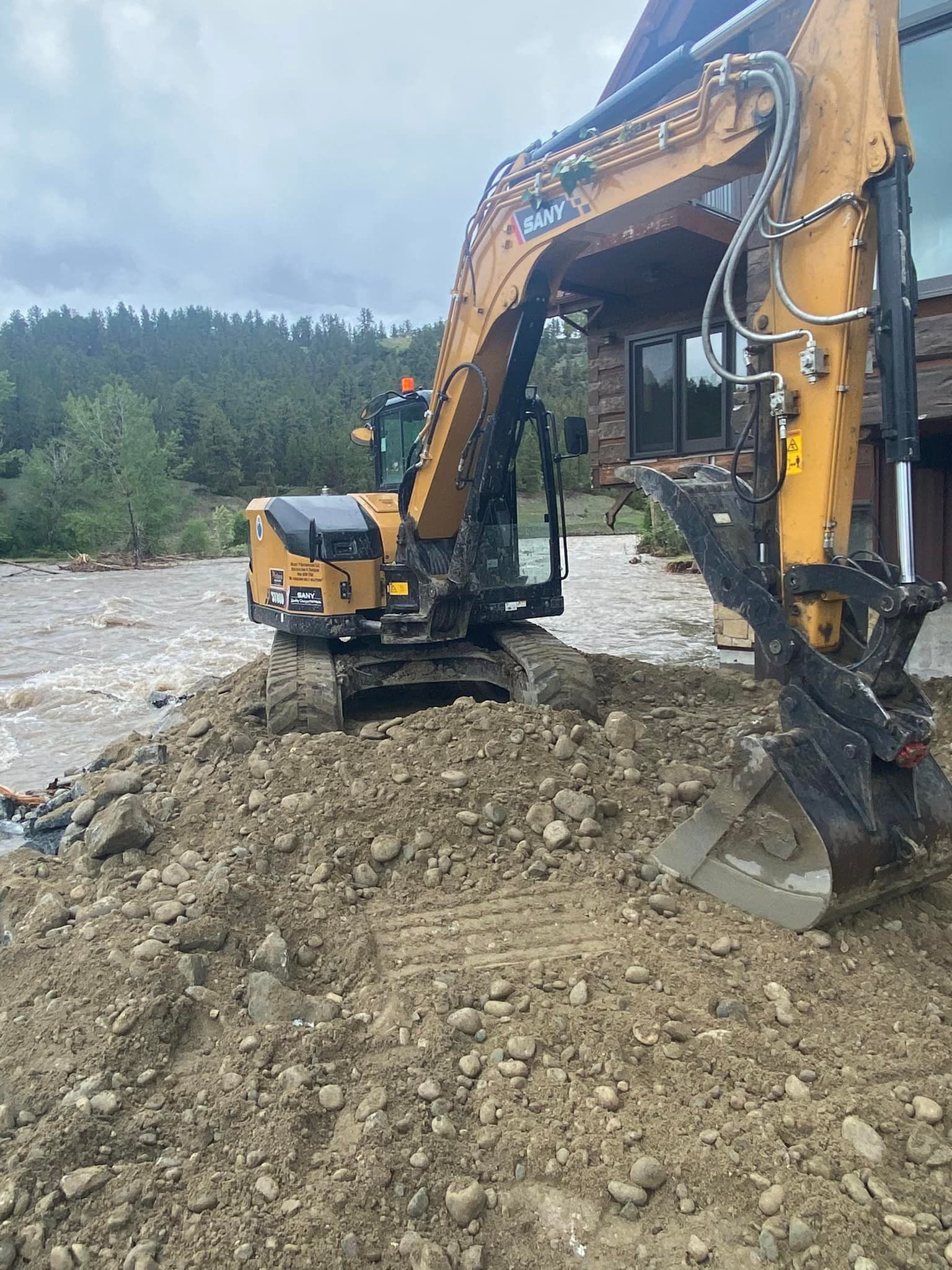 Yellow excavator moving earth near a flooded area, a person in green is in the cab, trees and a building are in the background.