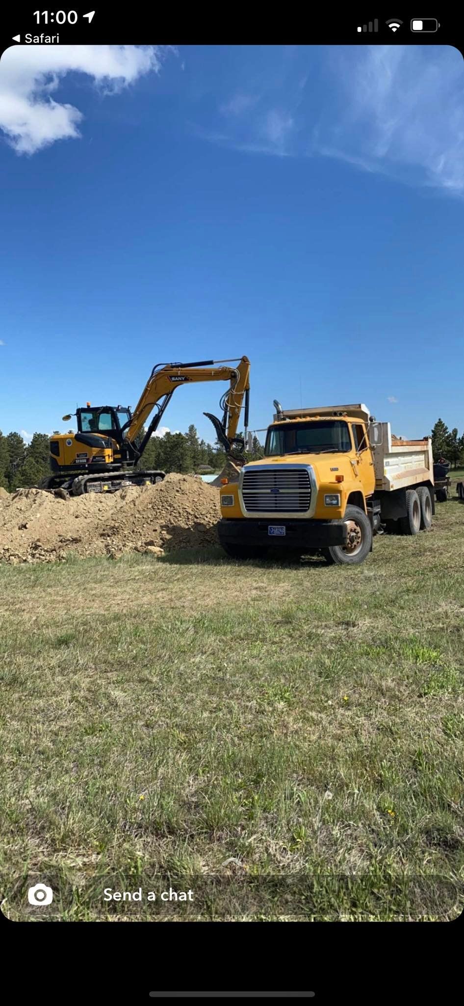 Yellow excavator loading dirt into a yellow dump truck in a grassy field under a blue sky.