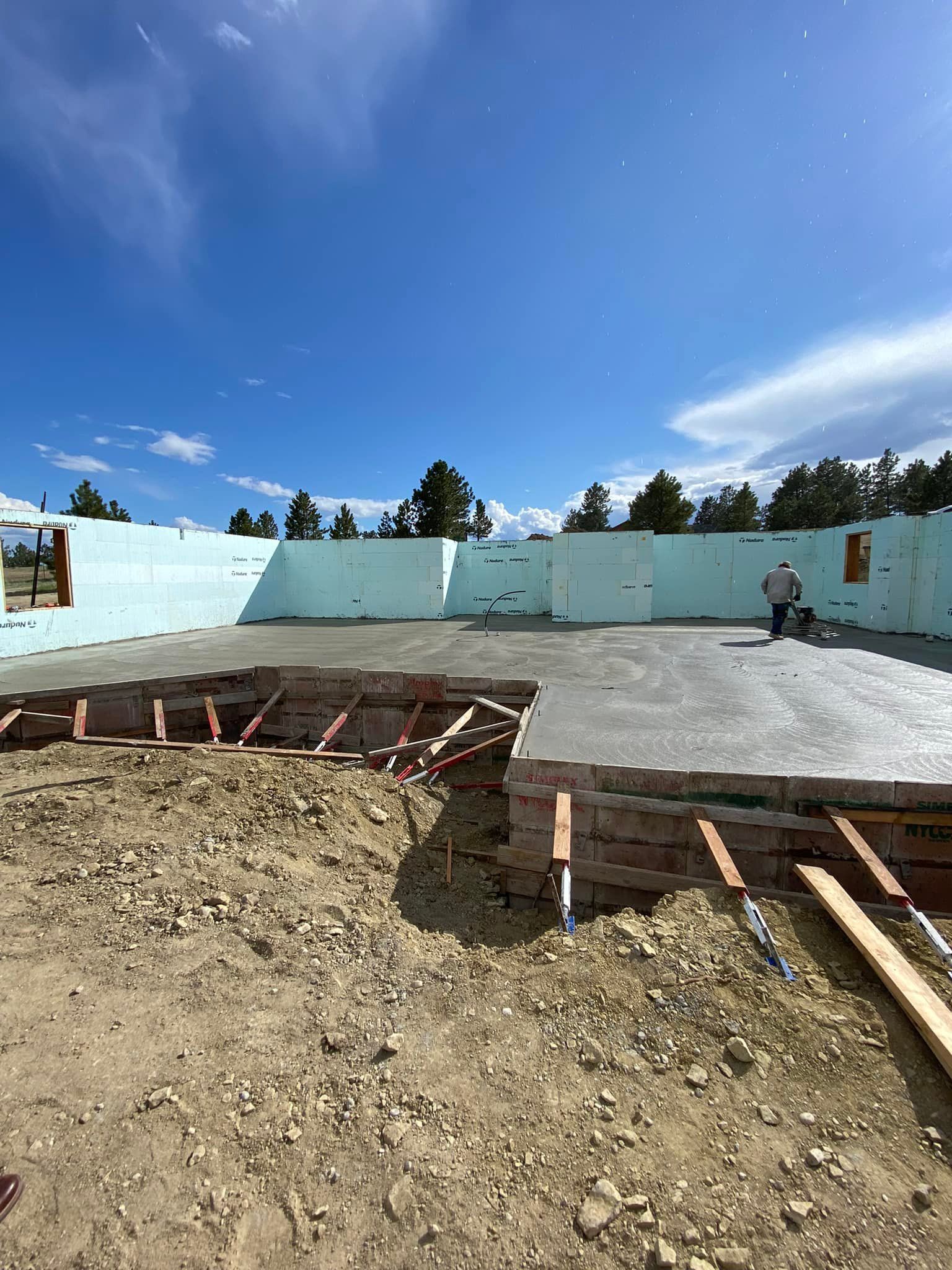 Construction site with concrete floor, blue insulated walls, wood framing, and a worker under a blue sky.