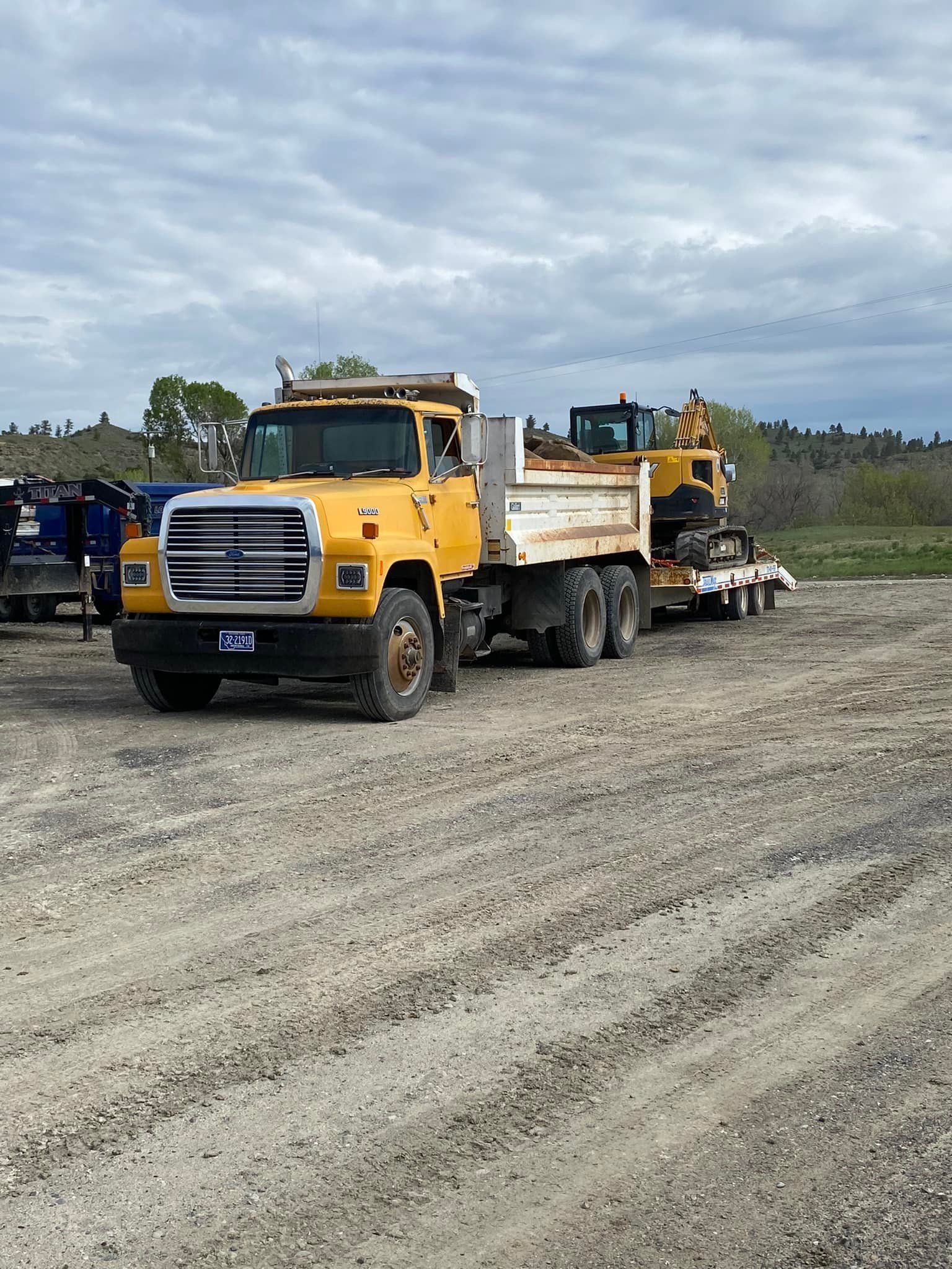 Yellow dump truck with a small construction machine on a trailer parked on gravel under a cloudy sky.