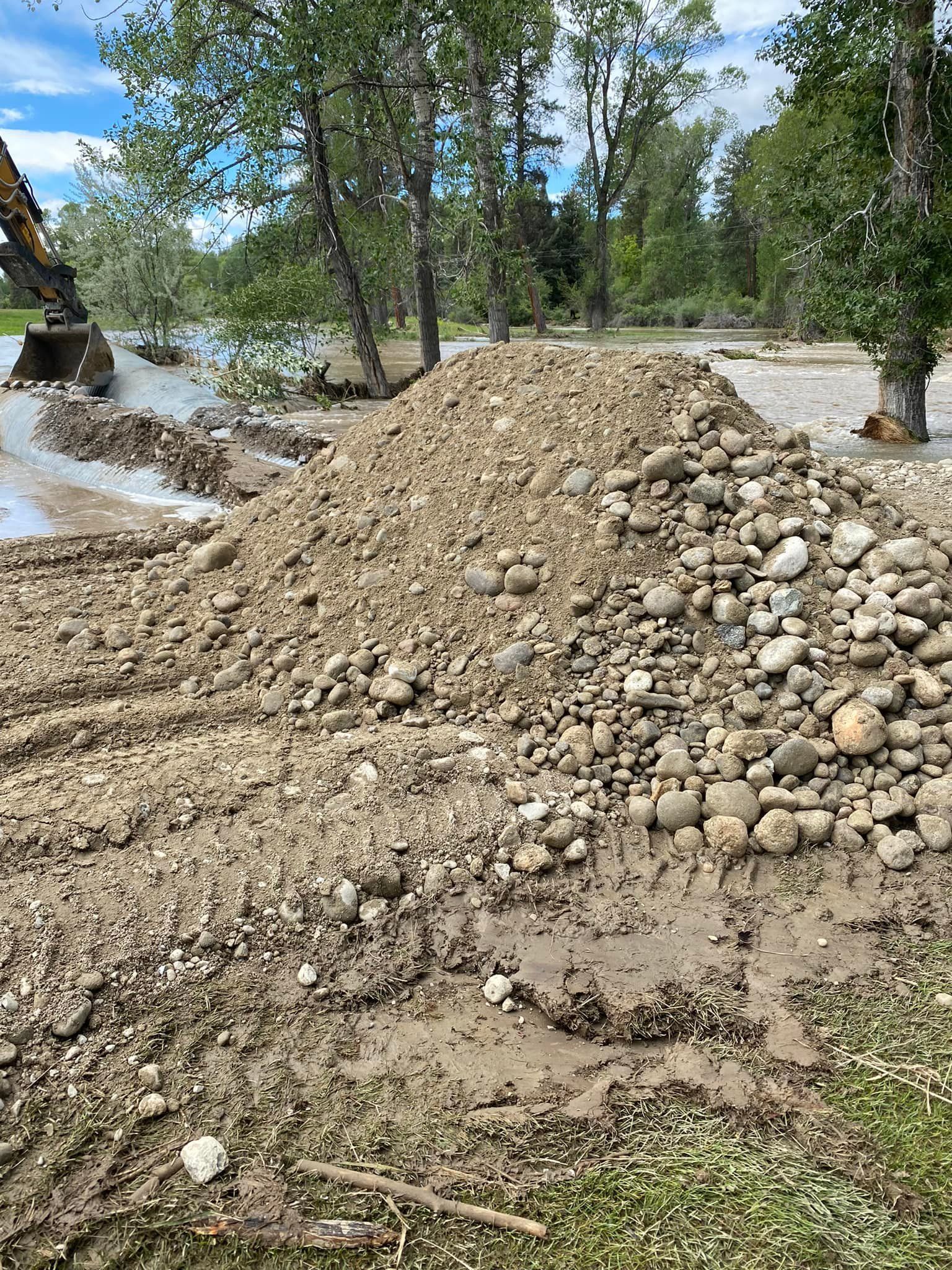 Pile of dirt and rocks next to muddy water, trees in the background, and an excavator.