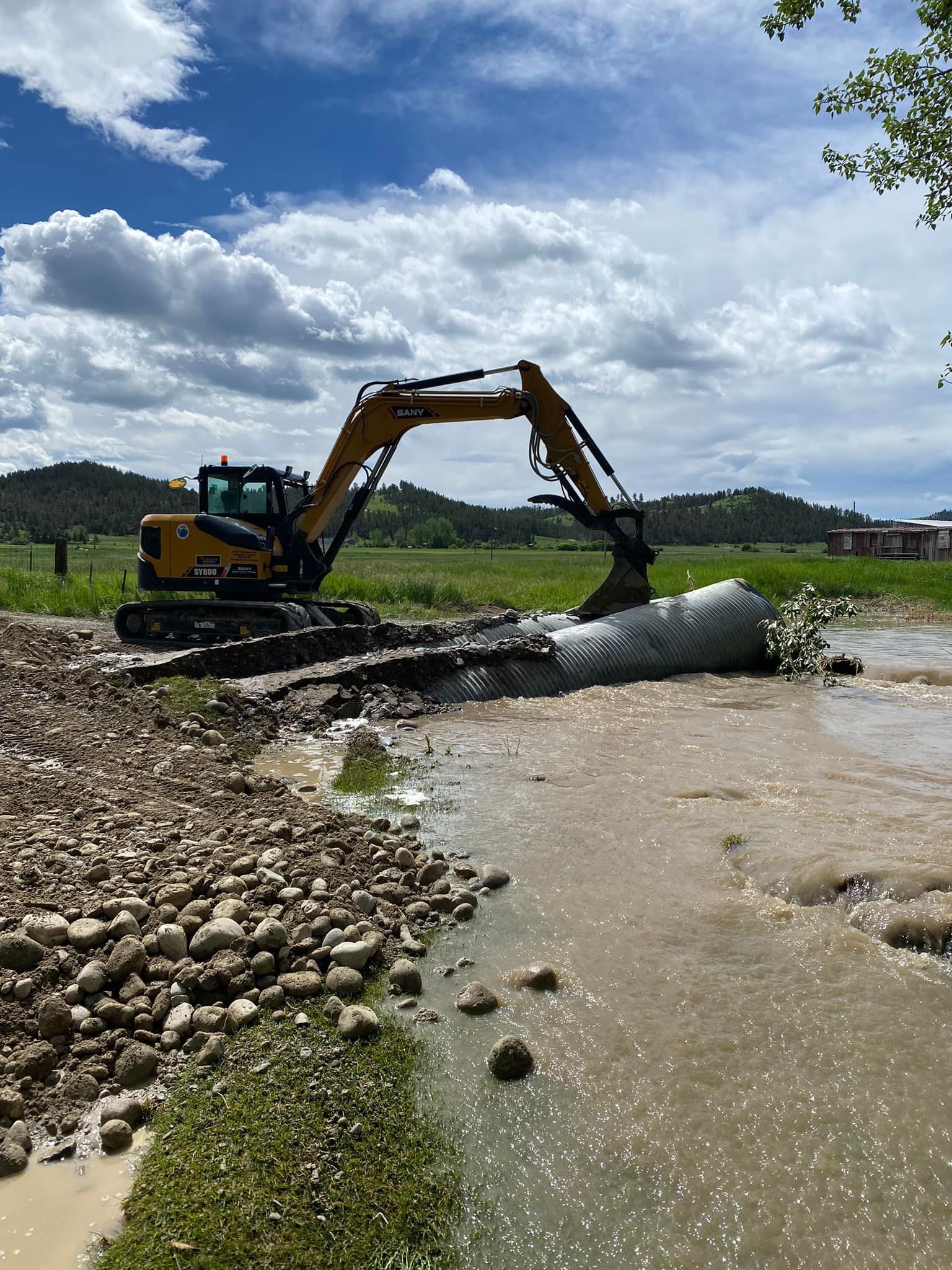 Excavator in muddy water, scooping material. Brown and tan tones, sunny day, field and distant hills.