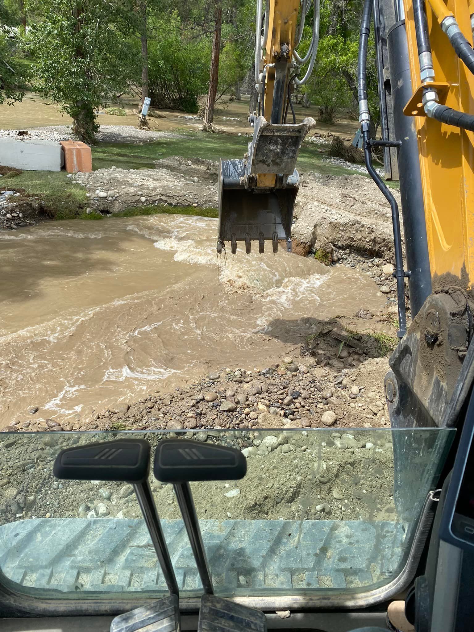 Excavator bucket scooping muddy water from a streambed; view from the cab.