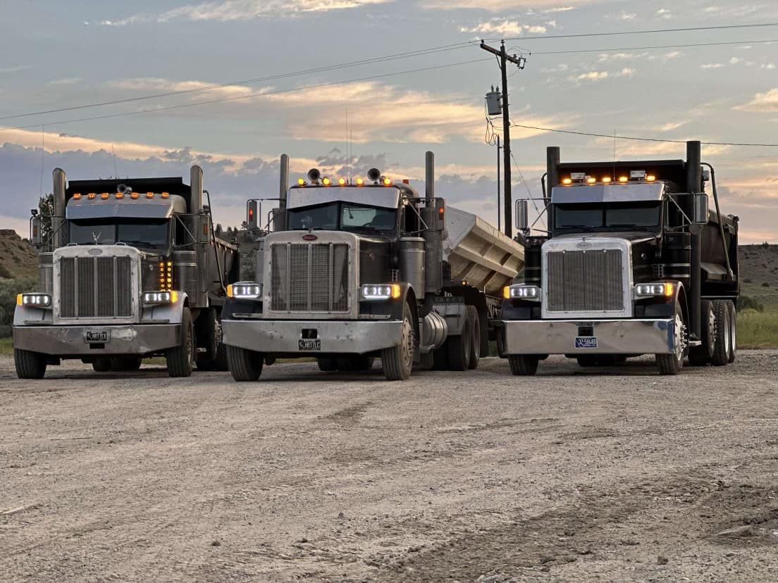 Three dump trucks parked on a dirt road, facing forward. Dusk sky.