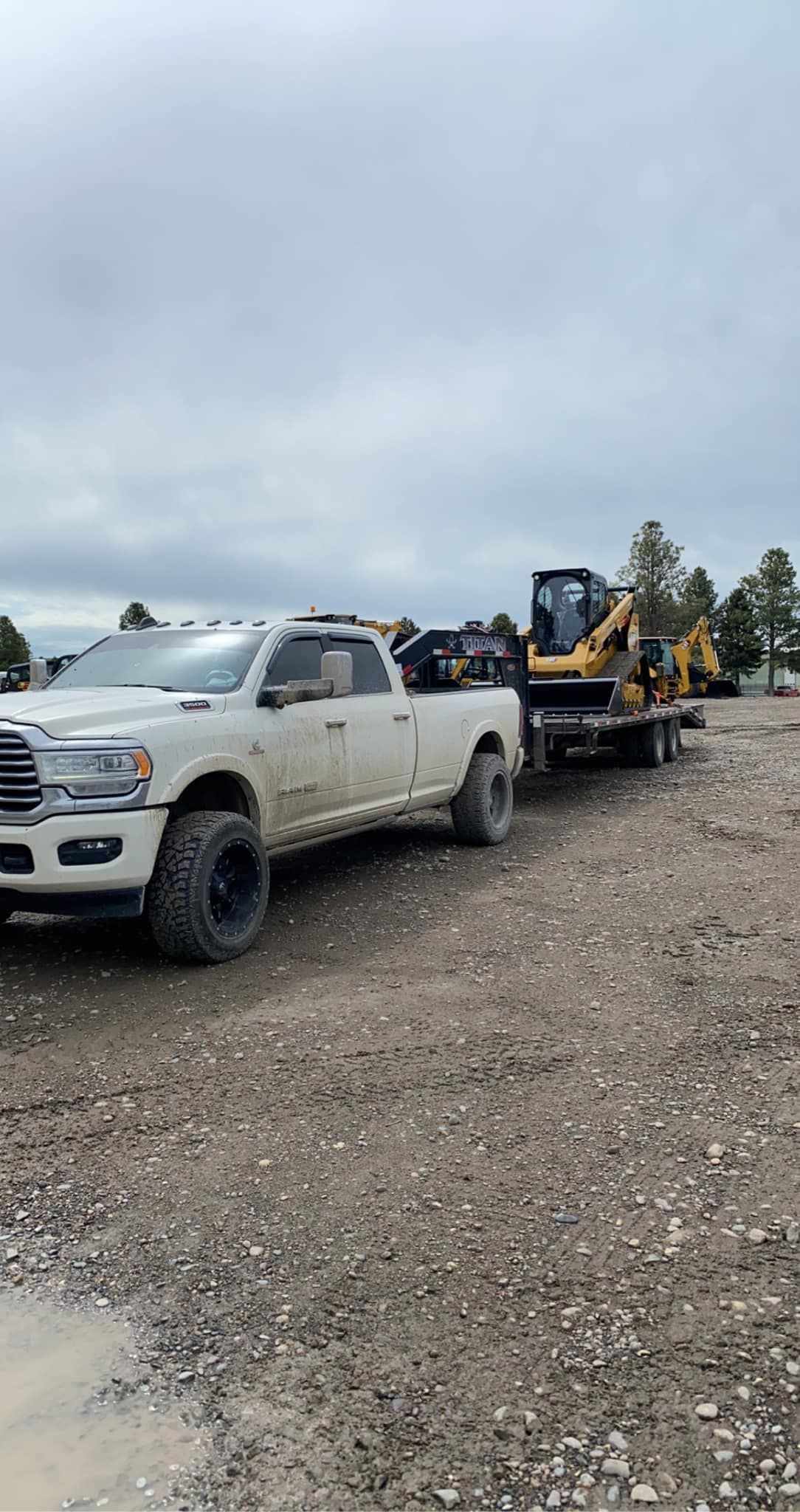 White truck towing a trailer with a small excavator on a muddy gravel road under a cloudy sky.