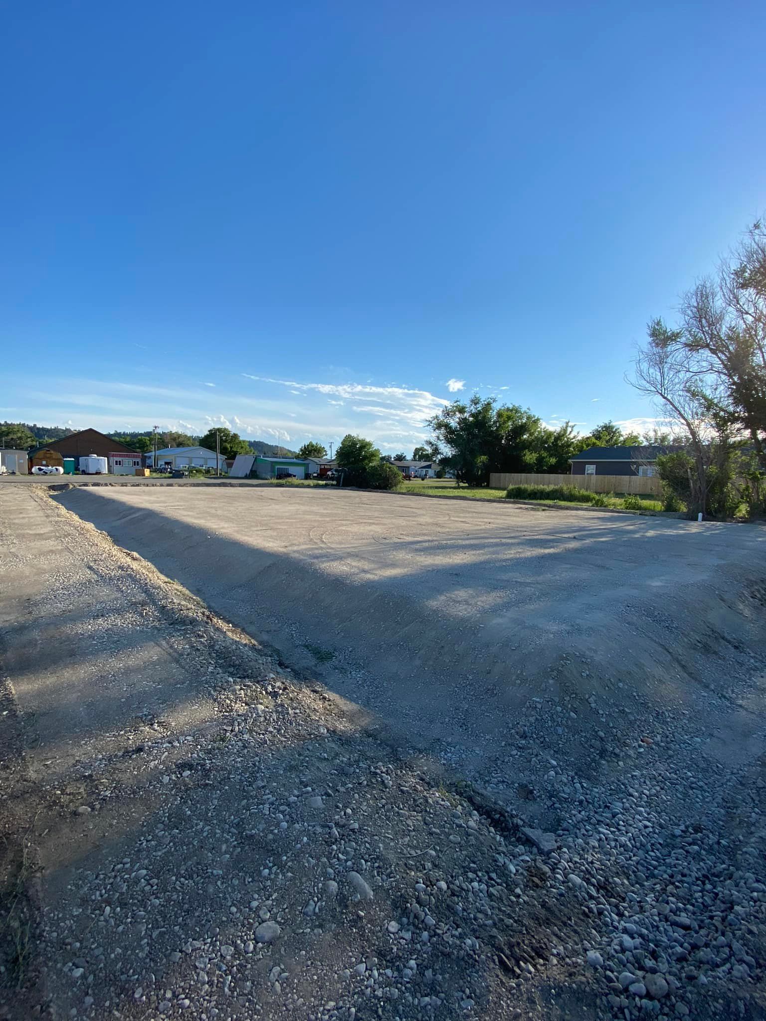 Empty gravel lot under a clear blue sky, buildings and trees in the background.