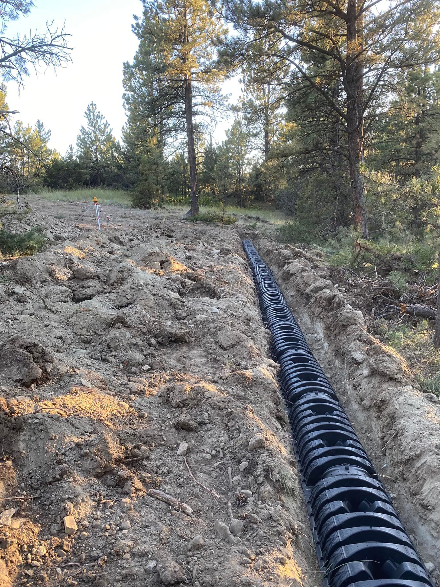 Black corrugated pipe laid in a trench in a dirt path in a wooded area.