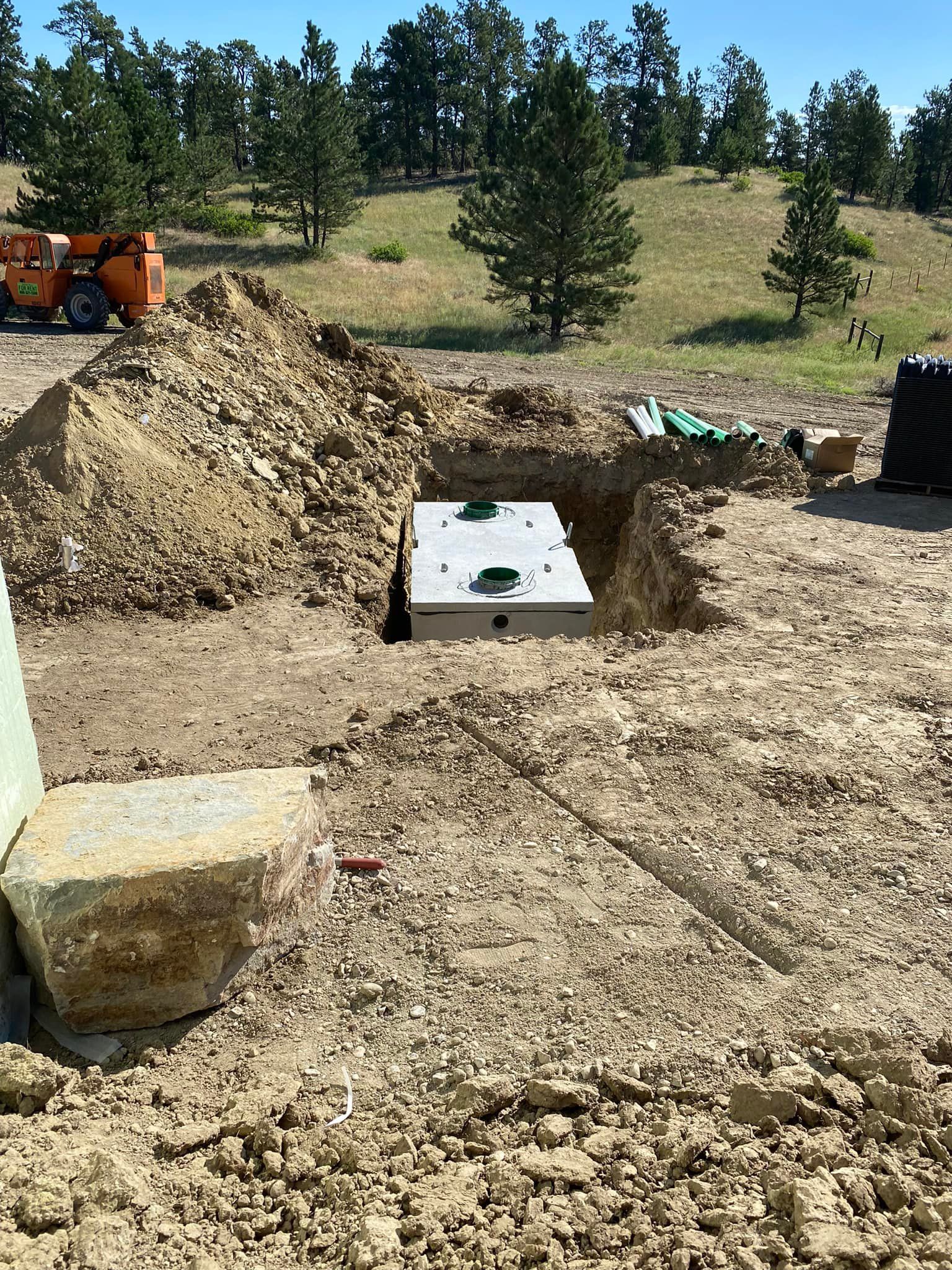 A rectangular septic tank partially buried in a dirt pit outdoors. Green pipes and an orange lift are visible.