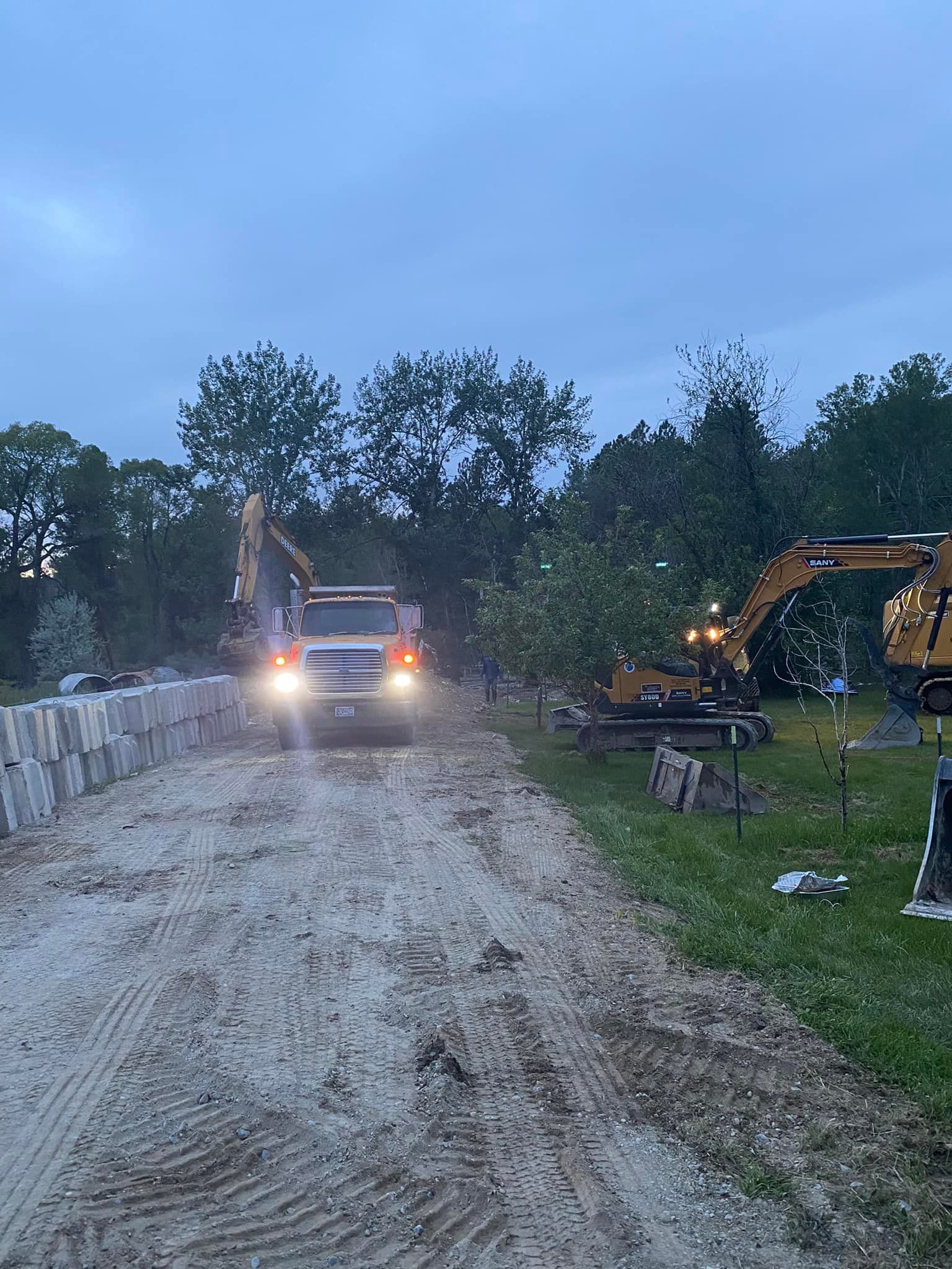 Truck and excavators on a dirt road, likely at a construction site. Gray sky, trees in the background.