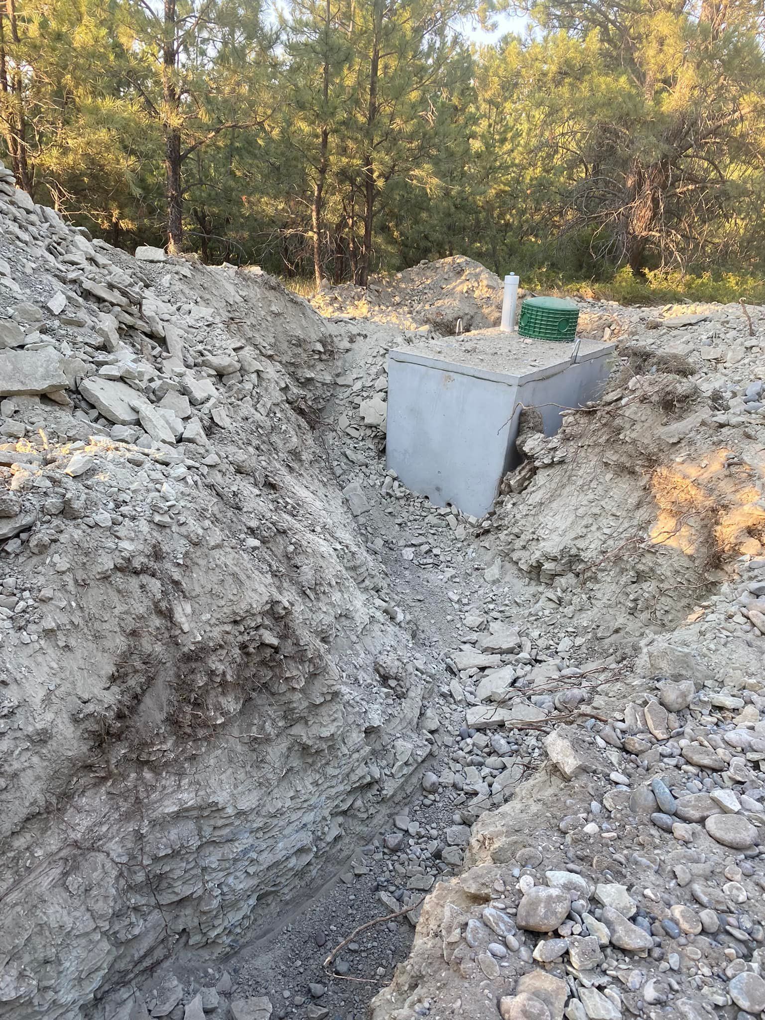 Construction site with a concrete structure and trench in front of trees.