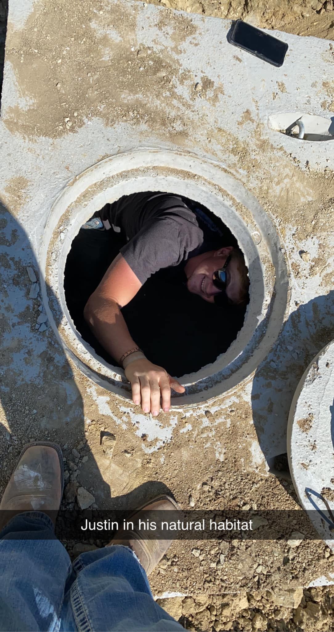 Man in a dark manhole, peering out. Gray concrete and dirt surround the opening.