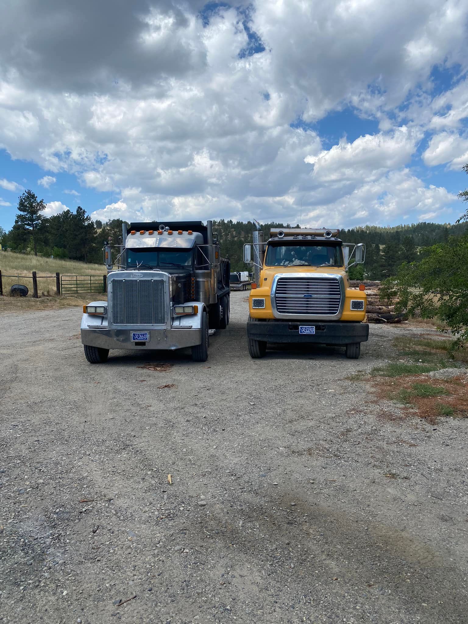 Two dump trucks parked on a gravel surface under a partly cloudy sky. One black, one yellow.
