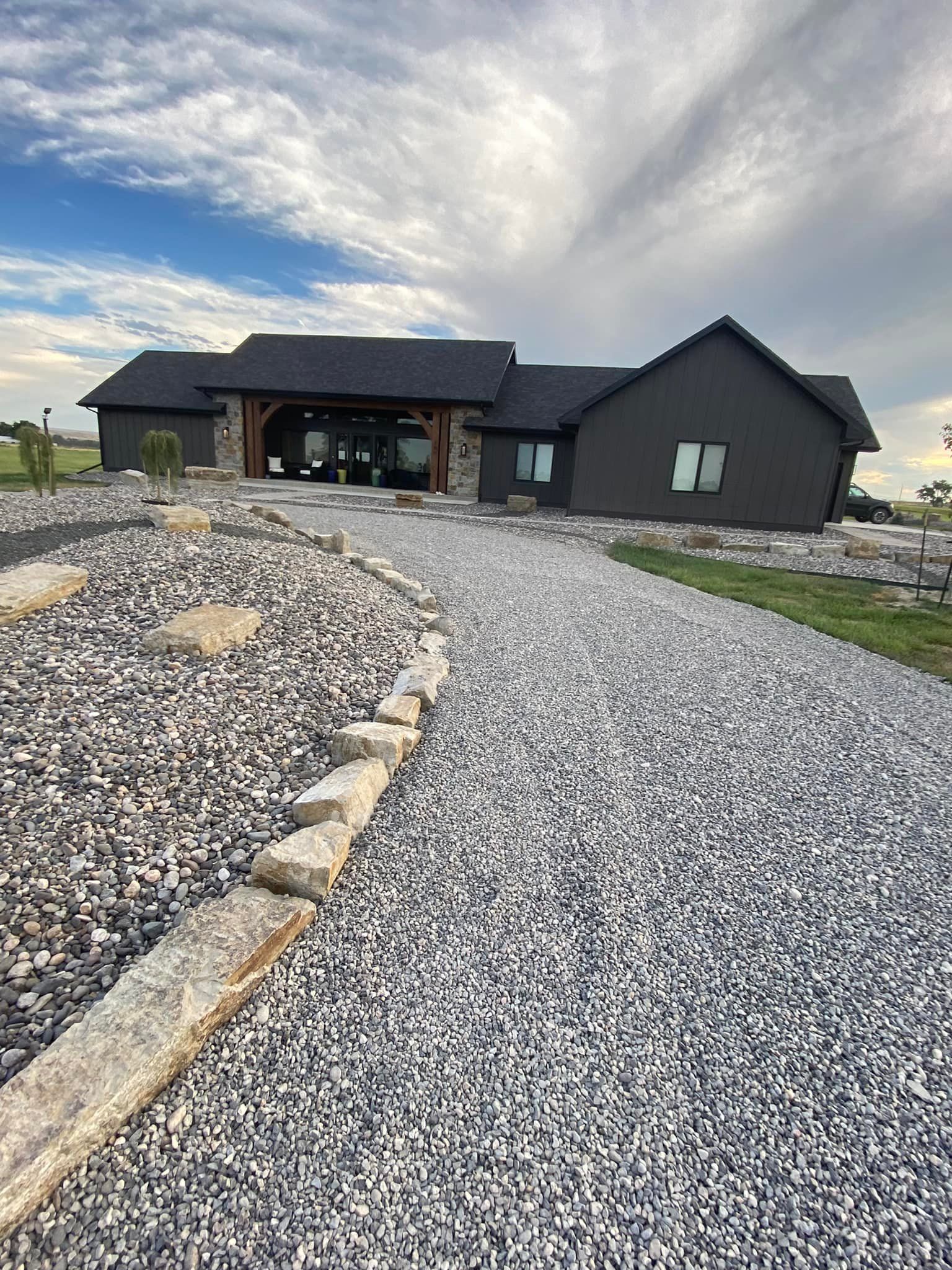 Gravel driveway leads to a modern, dark-gray house with stone accents and a cloudy sky.