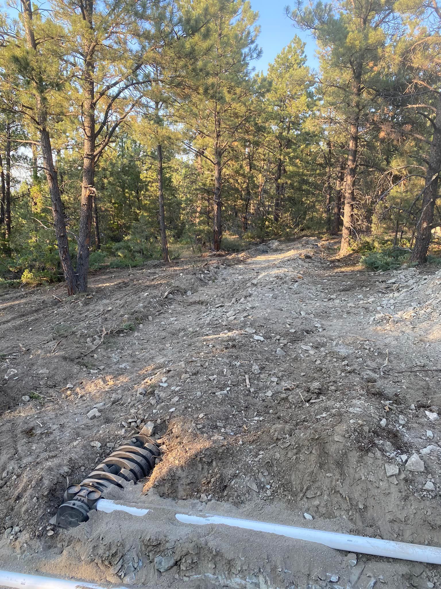 Piping and gravel in a dug-out area, with trees in the background, likely a construction site.