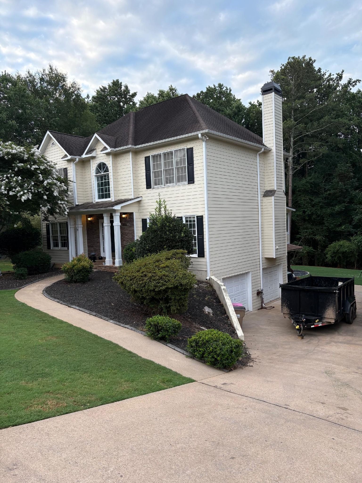 Beige house with black shutters and a winding walkway, set against a backdrop of green trees and cloudy sky.