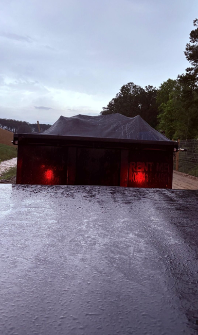 Rear view of a truck bed on a rainy day, tail lights illuminated, with a covered structure in the background.