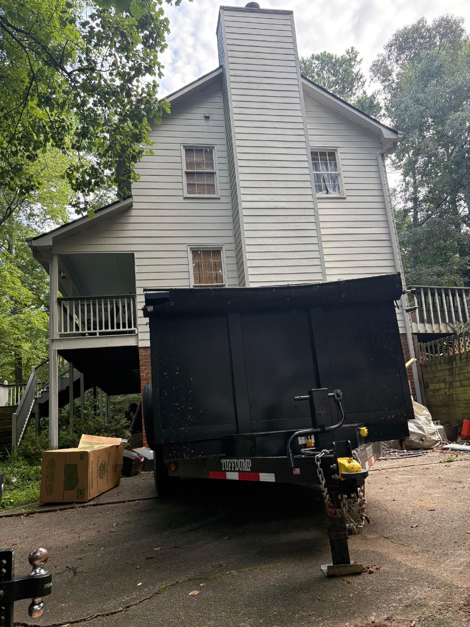 Black dumpster on a trailer parked in front of a two-story white house with chimney.