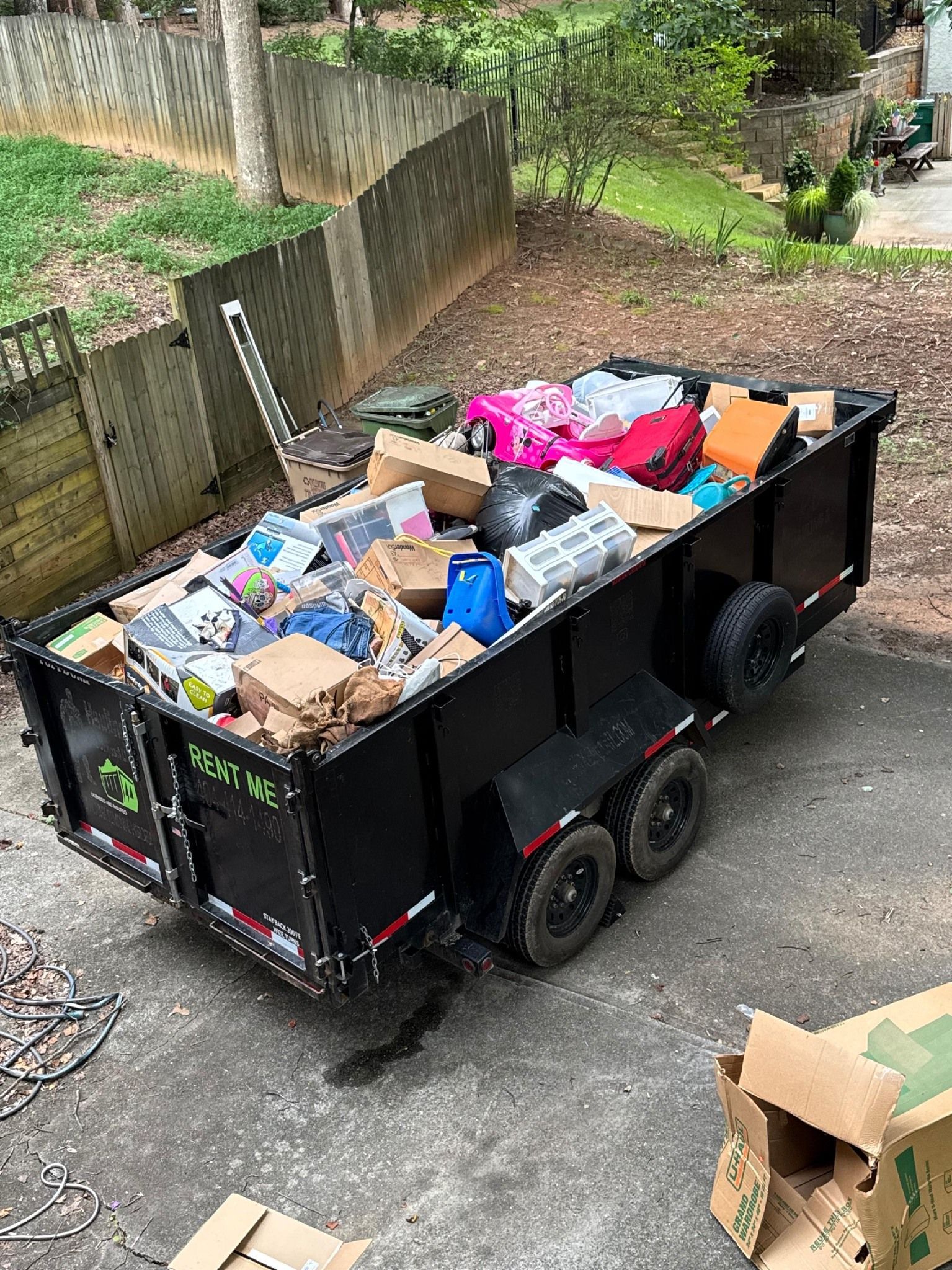 Black dumpster trailer overflowing with trash parked on a driveway, next to a fence and trees.