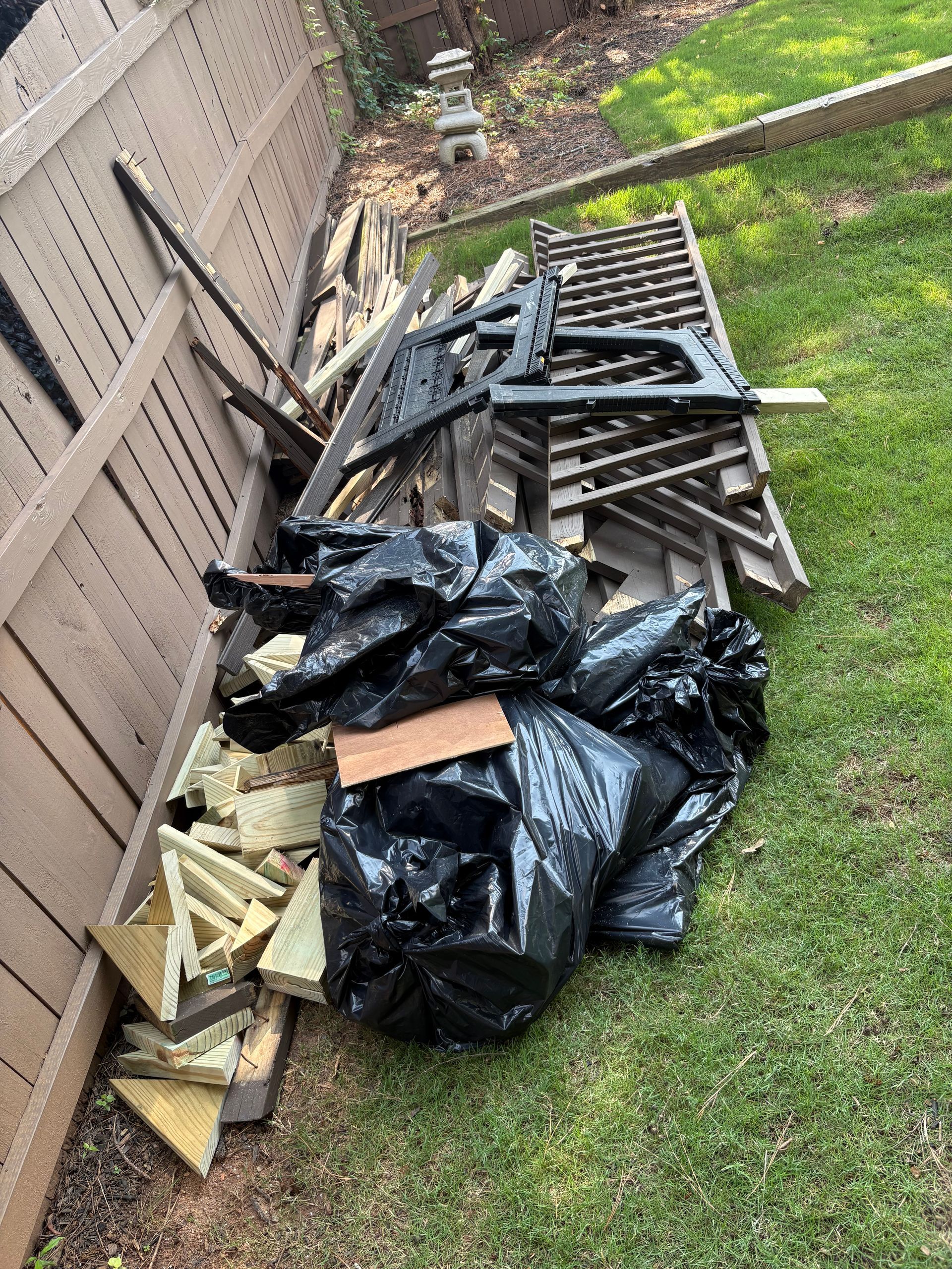 Pile of trash bags and construction debris next to a wooden fence and grass.
