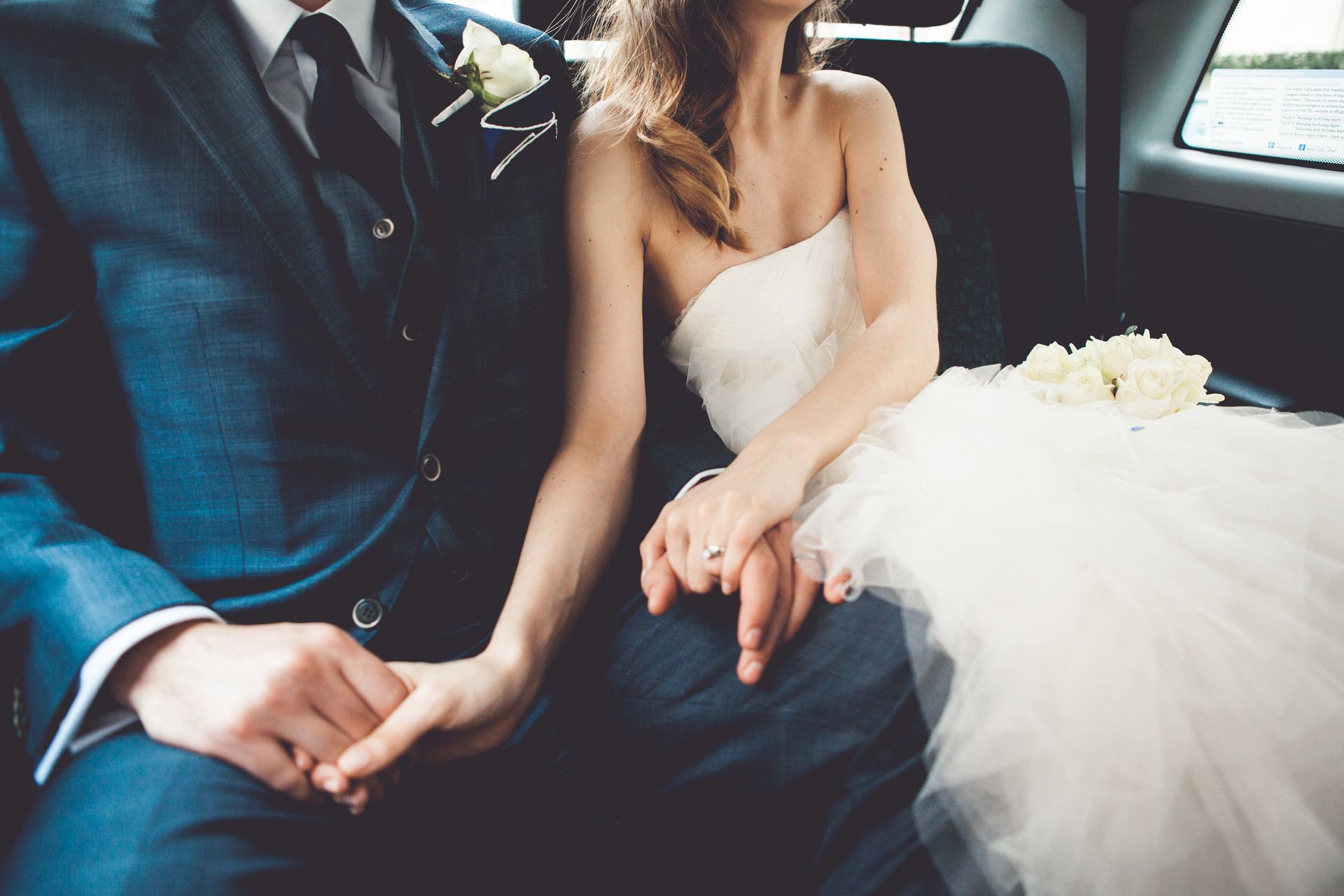 A bride and groom are holding hands in the back seat of a car.