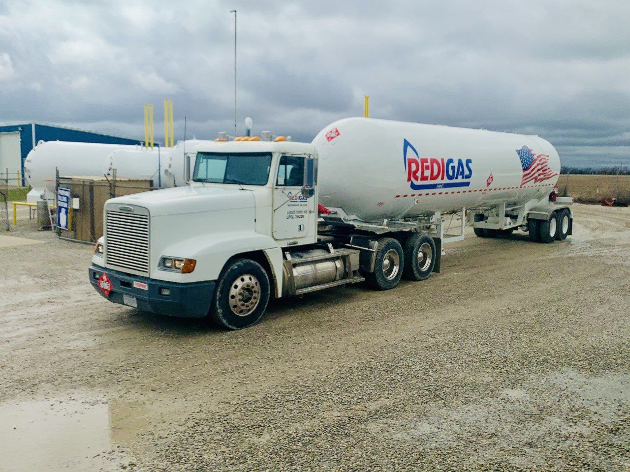 White Redigas tanker truck at a facility, hauling a large propane tank. Overcast sky.