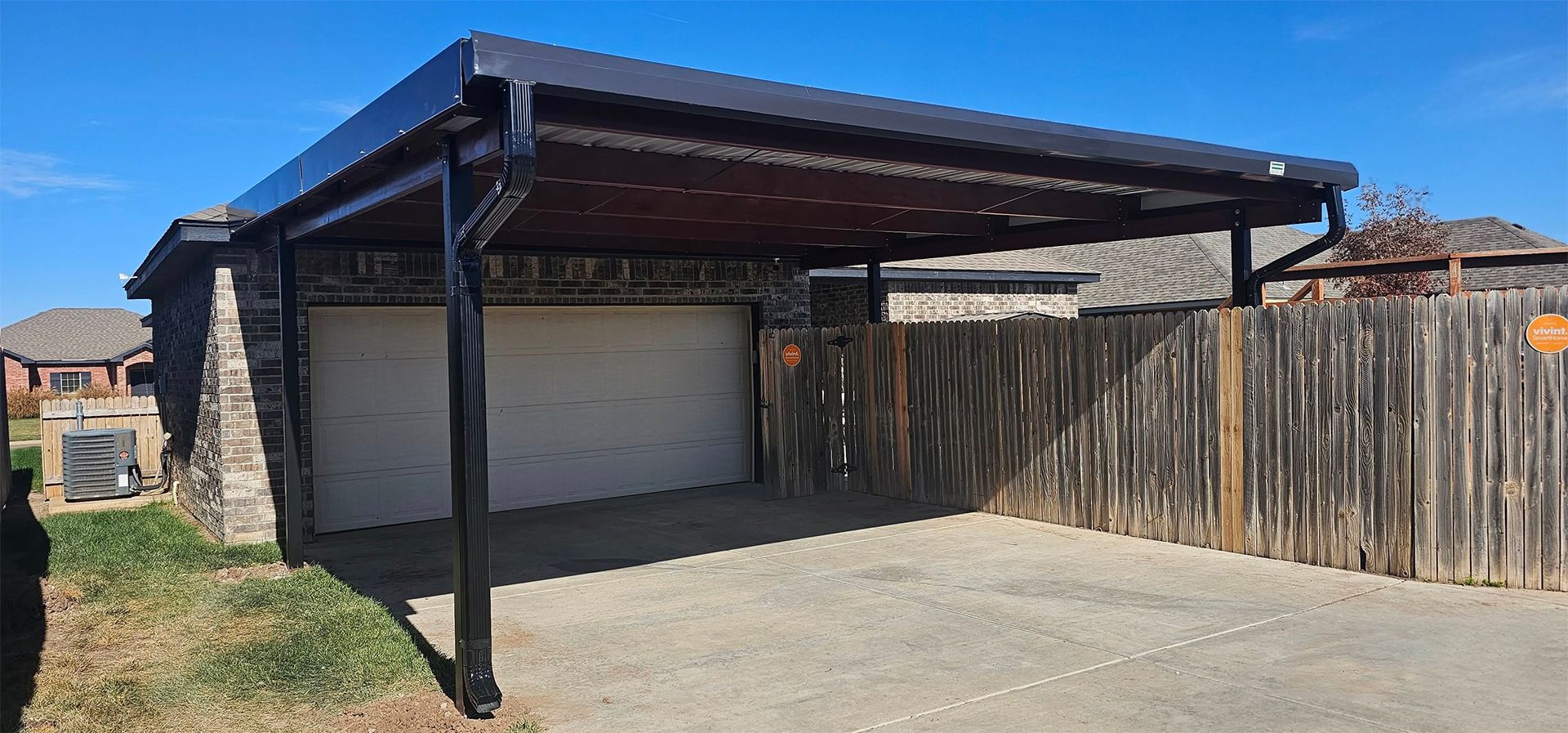 Carport over a garage. Black frame and roof, brick wall, gray concrete driveway, wooden fence.