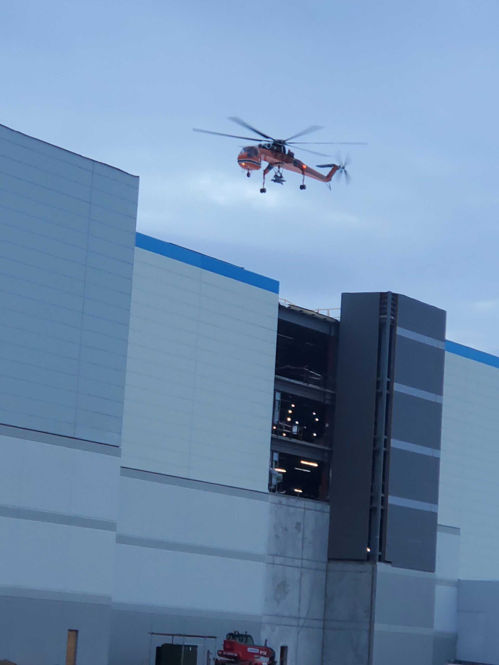 Orange helicopter flying near a large industrial building on a cloudy day.