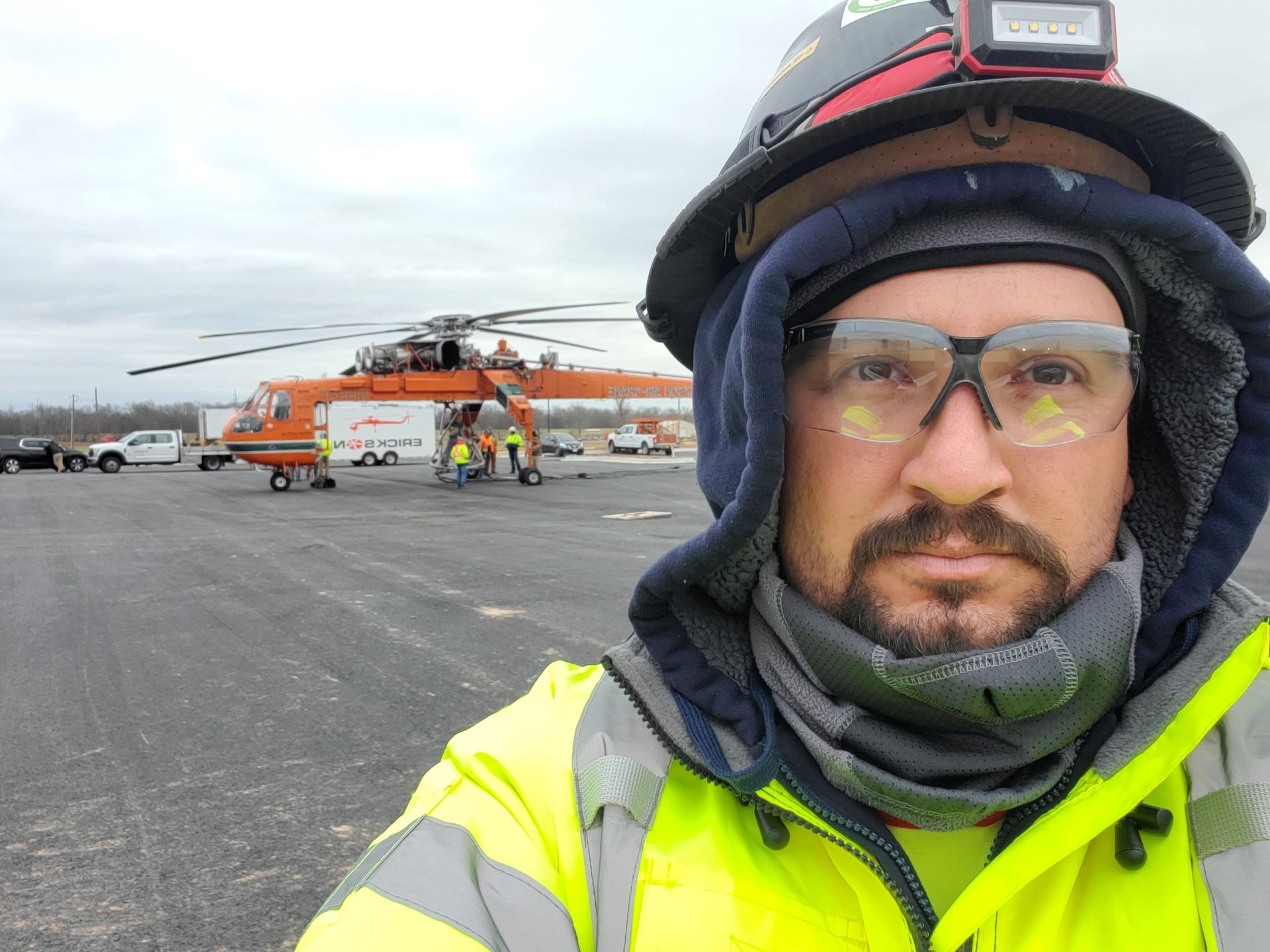 Man in work gear takes selfie in front of large orange helicopter on tarmac. Overcast day.