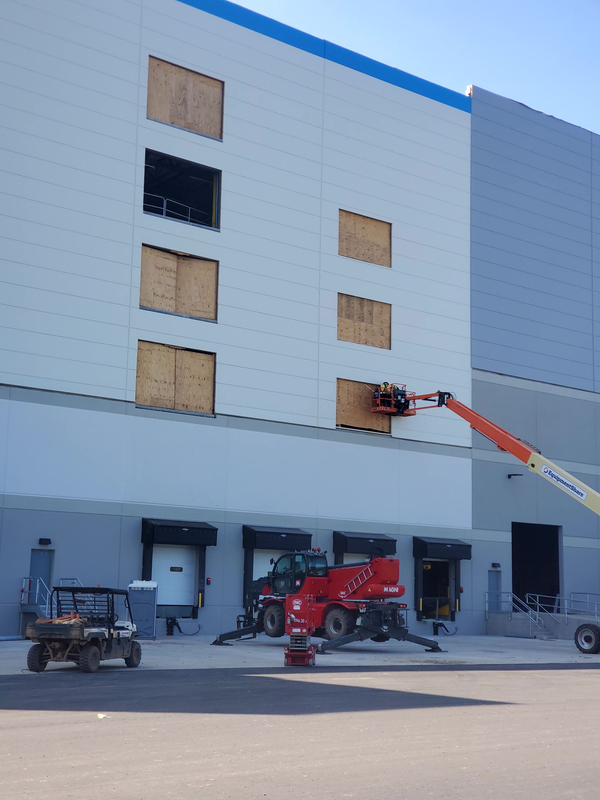 Construction on a white warehouse, plywood over window openings. A lift with worker in use. Vehicles in front.