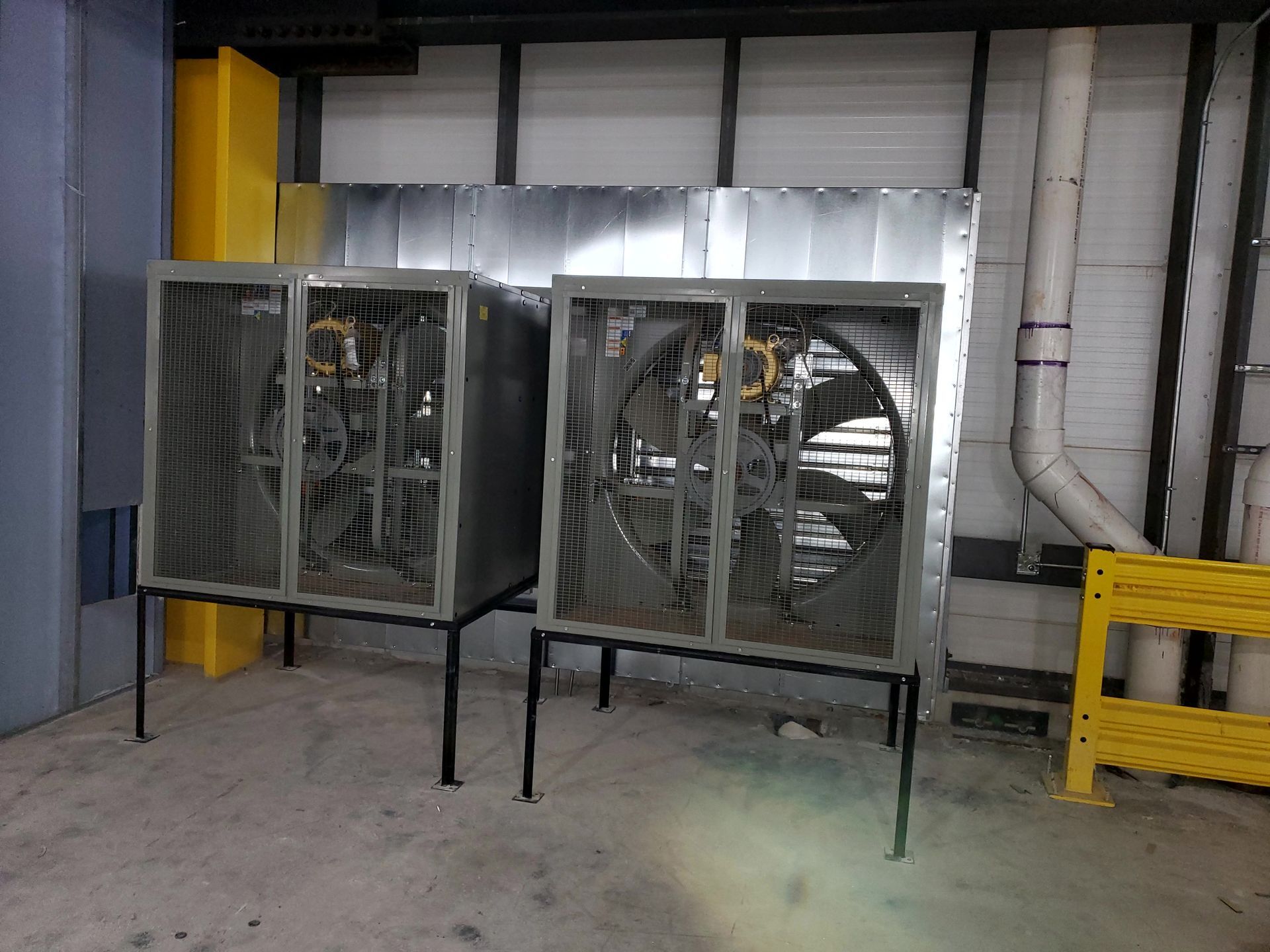 Two industrial fans, metallic with protective grates, on black legs. Pipes and yellow safety barrier in background.