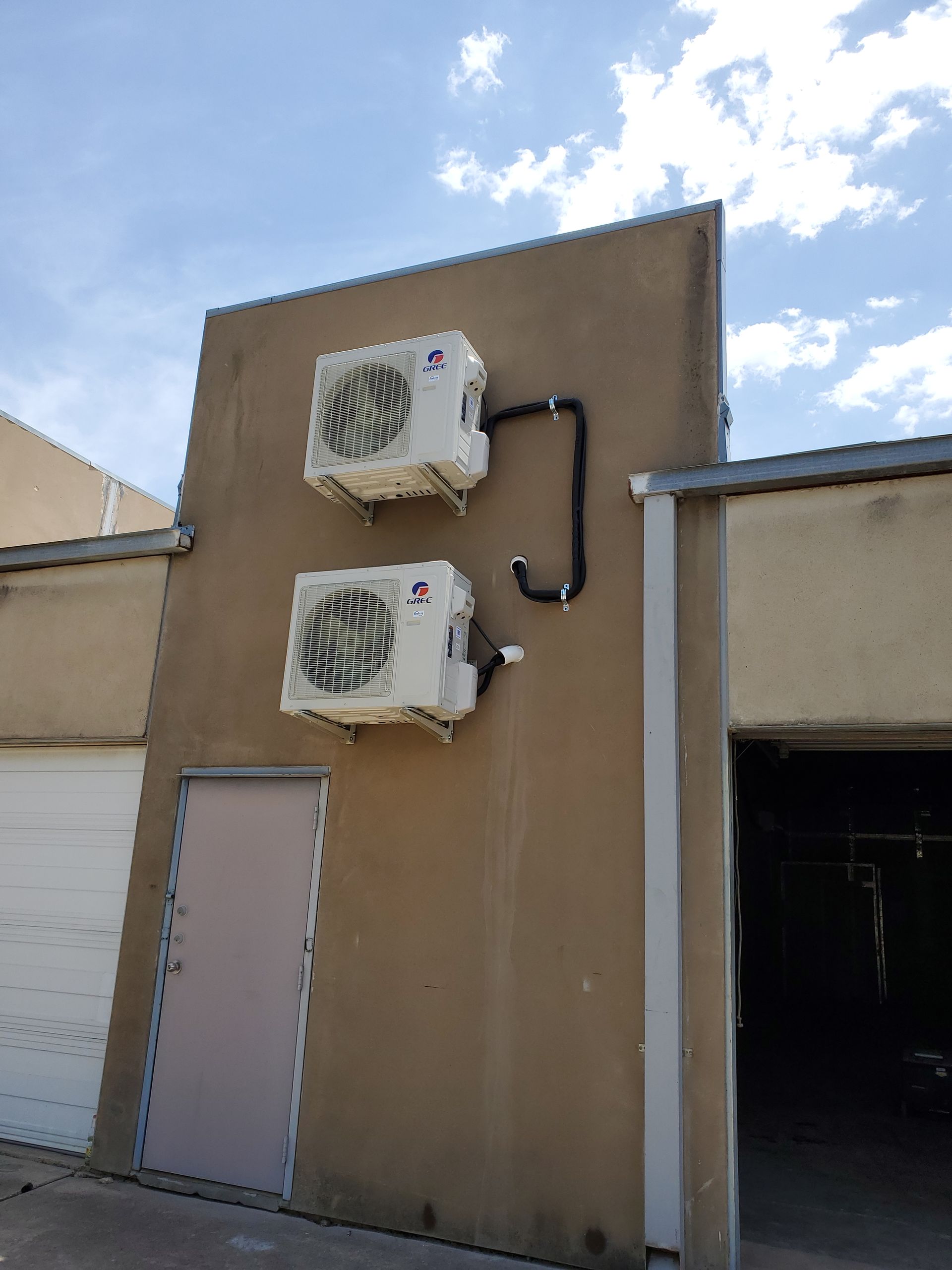 Two air conditioning units mounted on a beige building wall with black pipes and a door below.