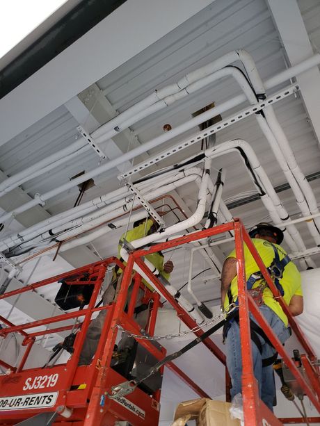 Construction workers on a lift installing pipes on a white ceiling. They wear safety vests and harnesses.
