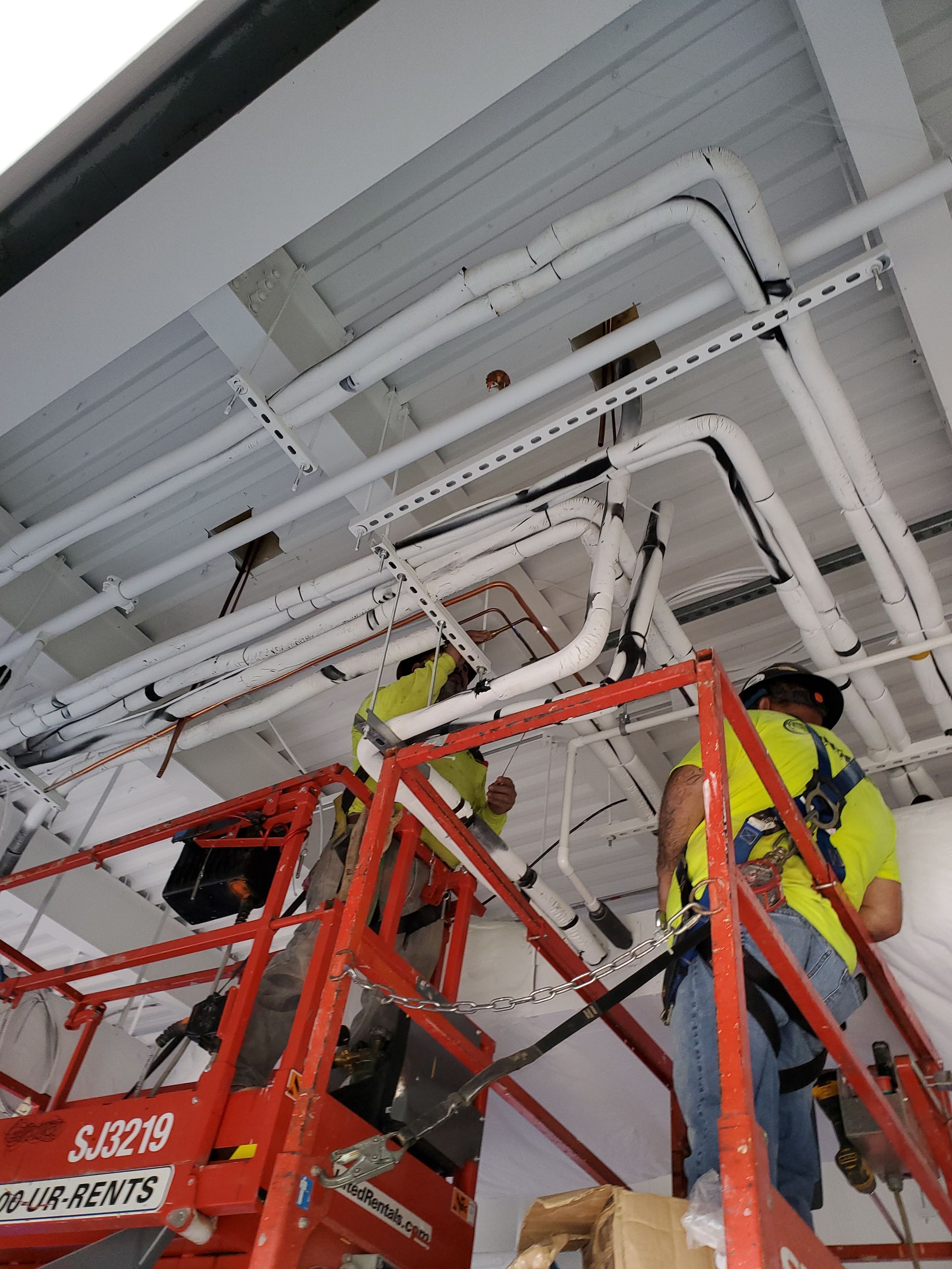 Two workers in a lift install pipes on a white ceiling. They wear safety vests, and the pipes are white.