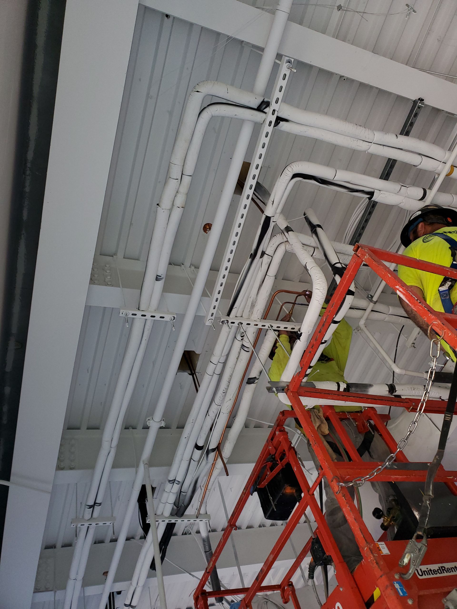 Workers on a lift installing white pipes along the ceiling of a building.