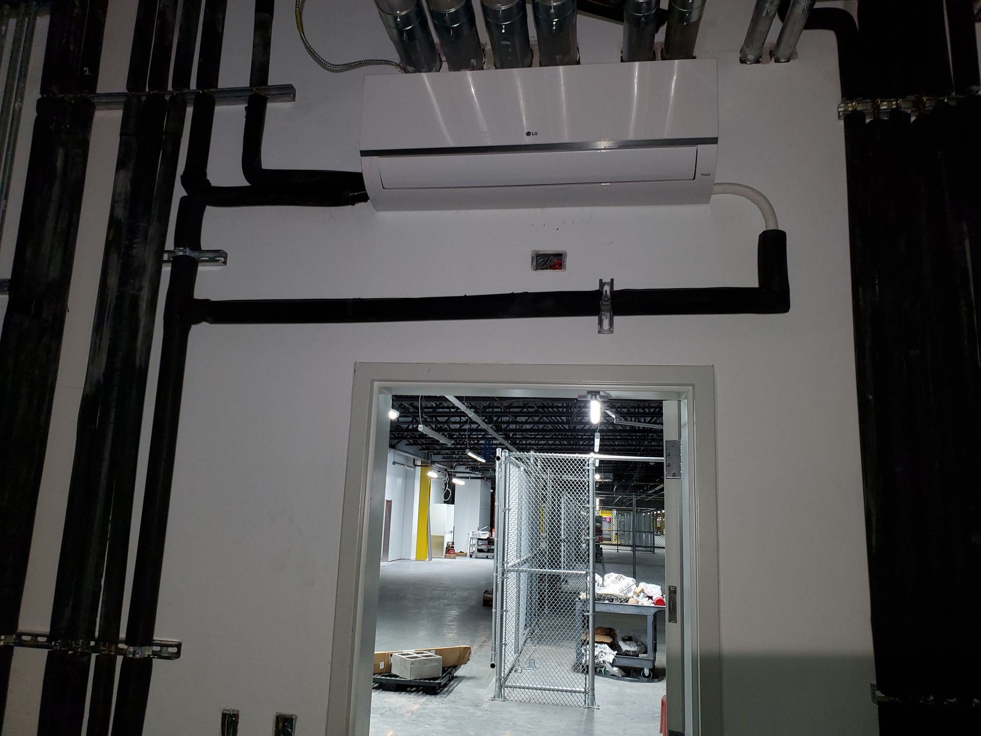 White wall with an AC unit above a doorway, black pipes, and a glimpse into a warehouse.