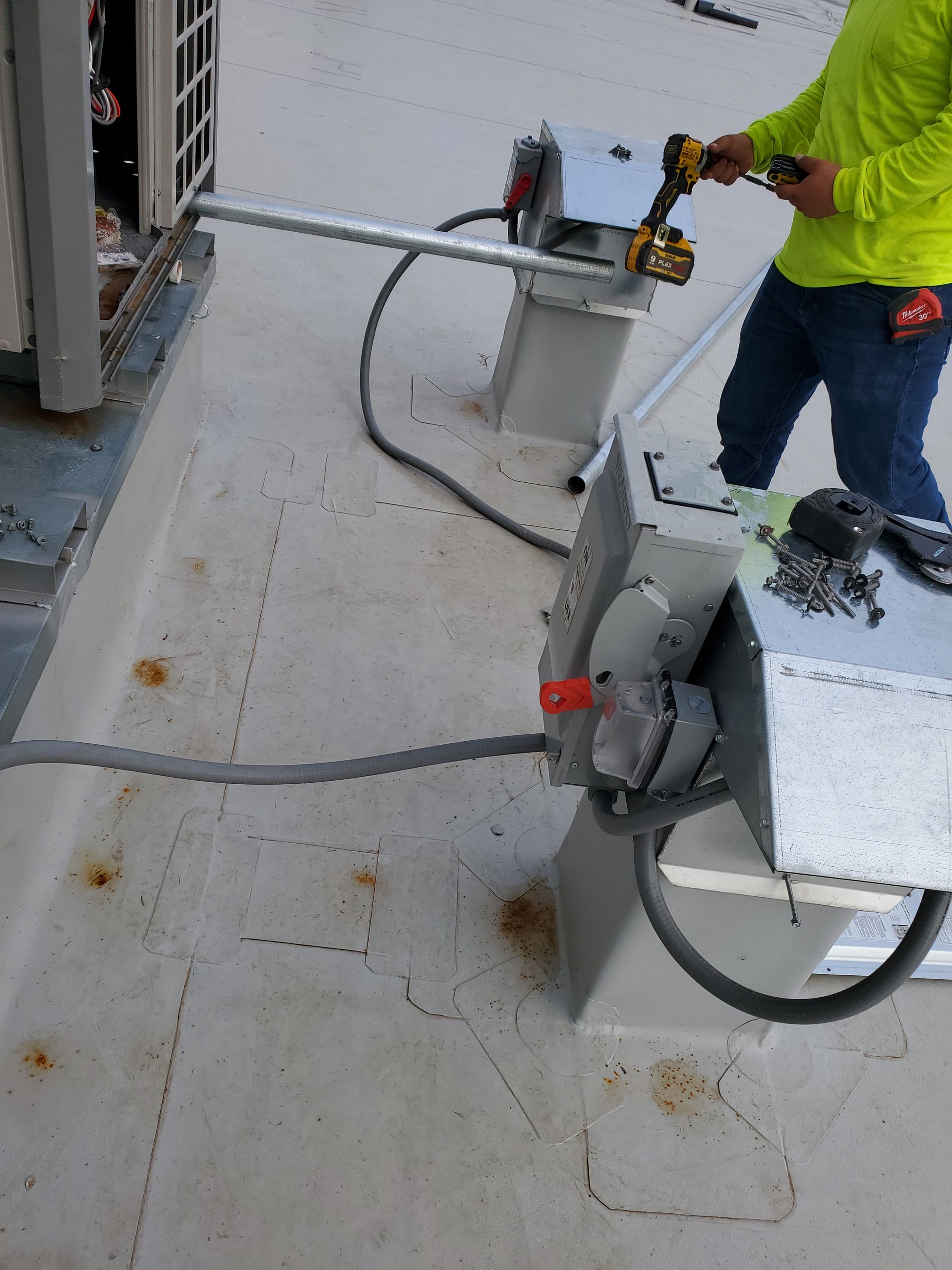 Person in yellow shirt using a drill on rooftop electrical equipment.
