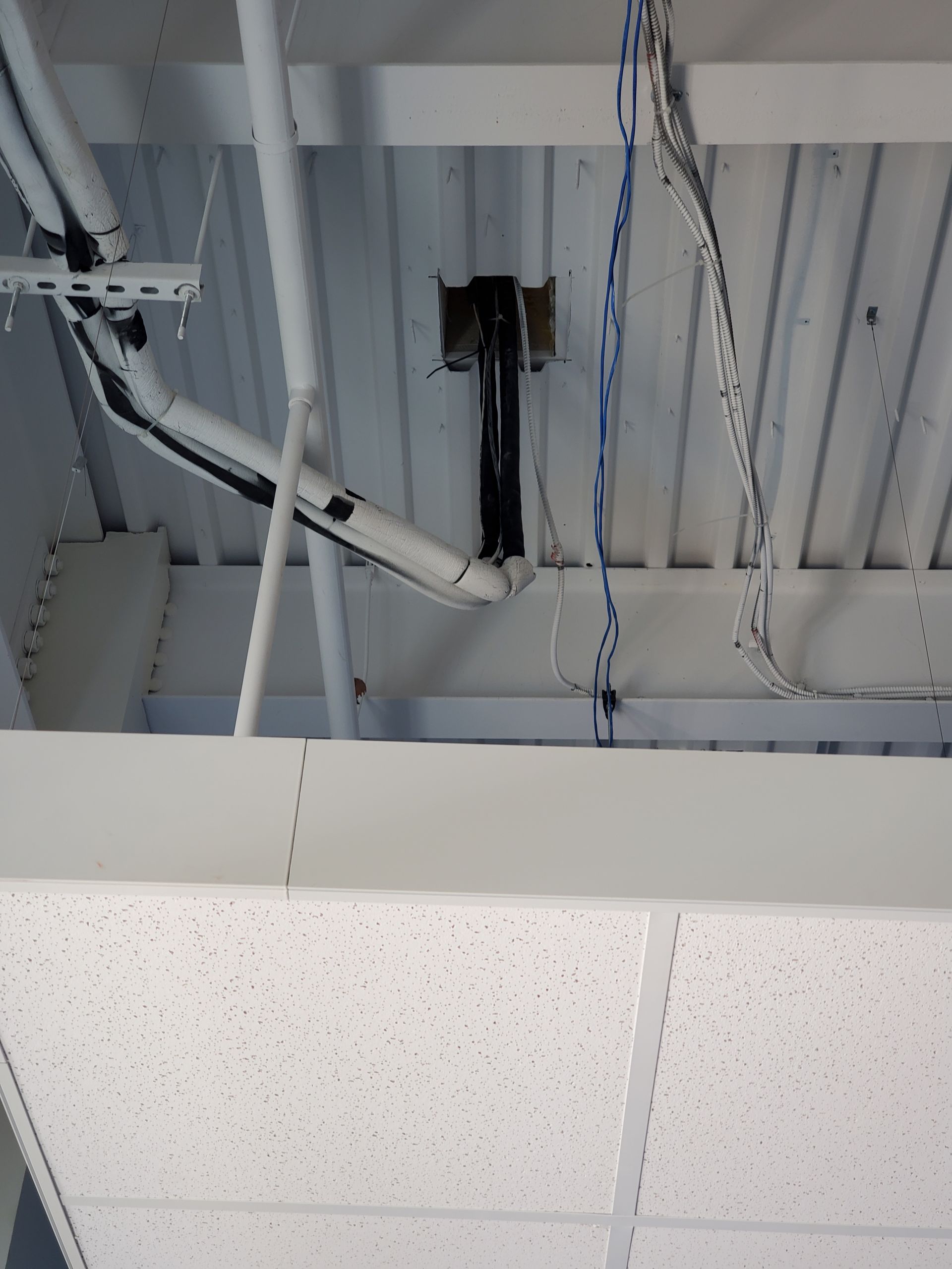Overhead view of a white ceiling with exposed pipes, wires, and a black projector mount.