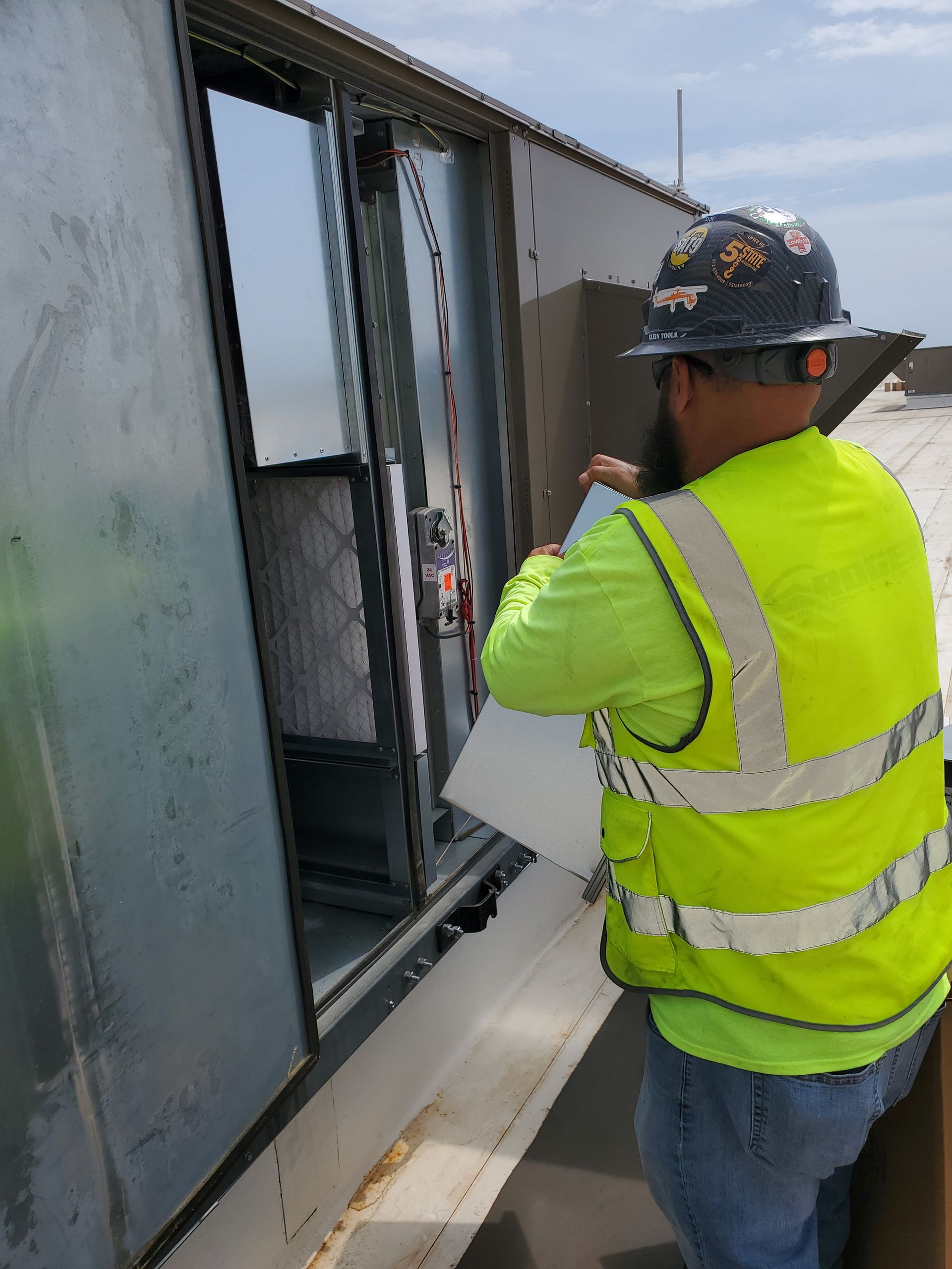 Man in safety vest replacing air filter on rooftop HVAC unit.