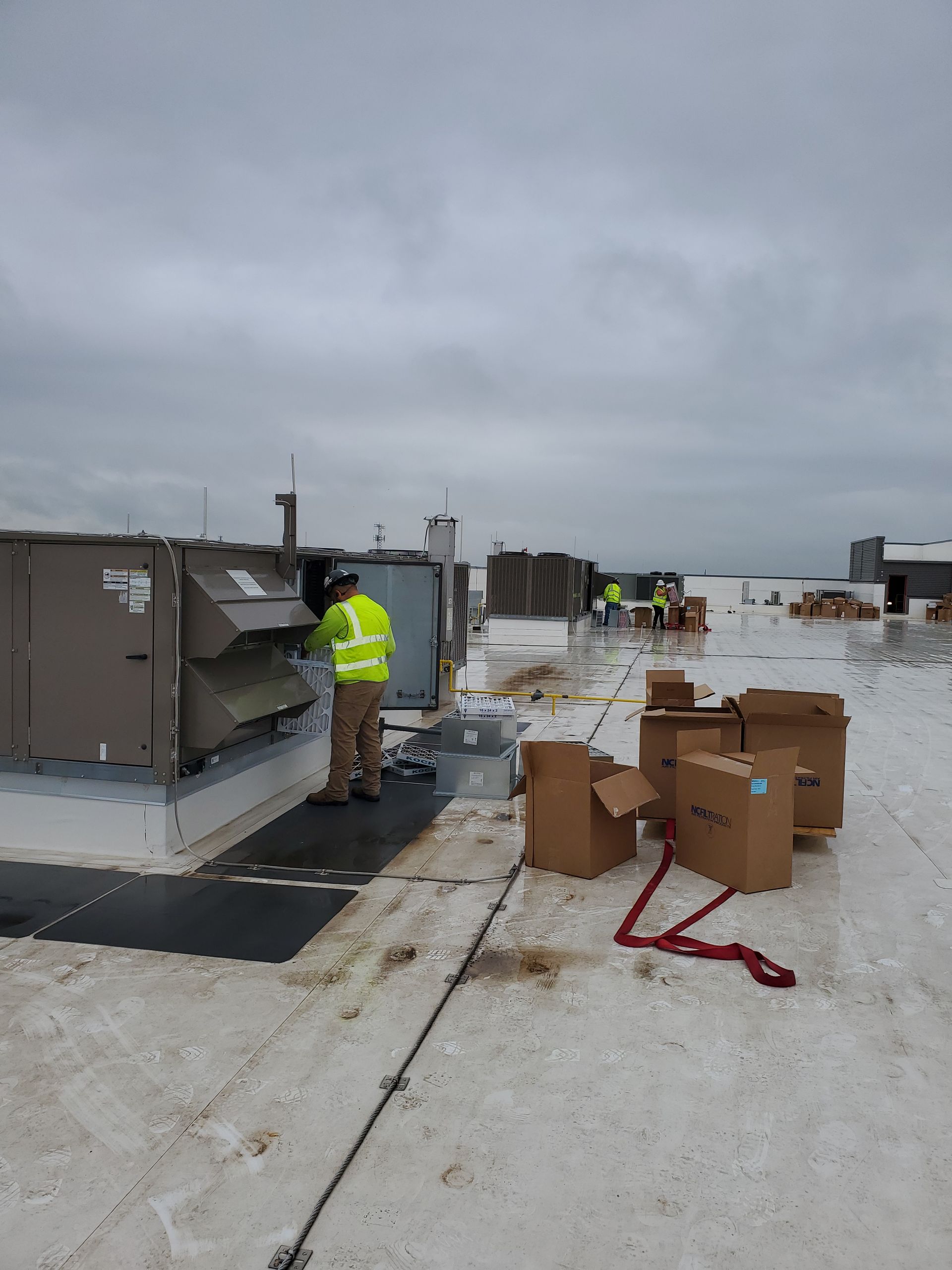 Workers in safety vests on a rooftop, near HVAC equipment. Cloudy sky, boxes, and tie-down straps are present.