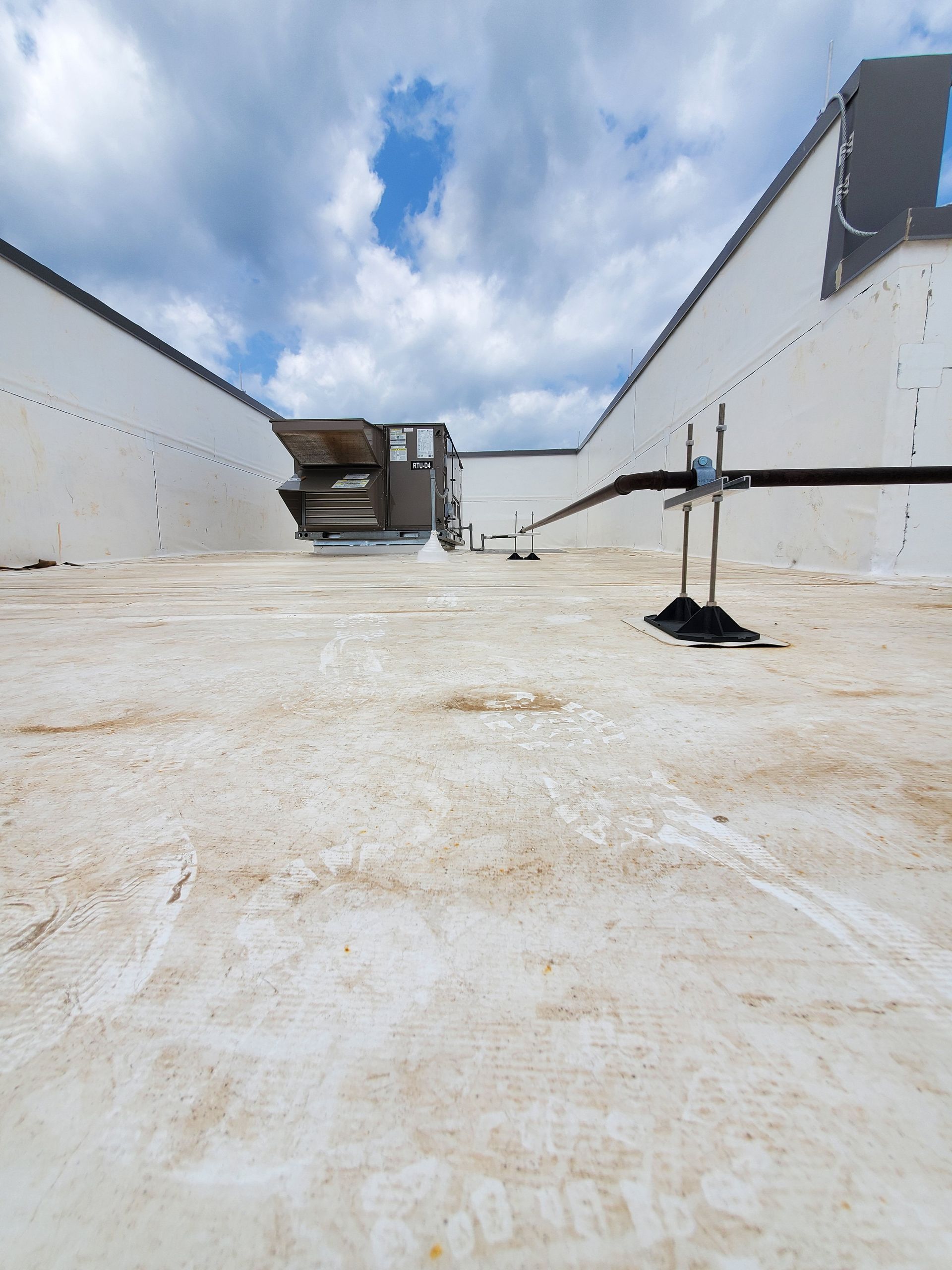 White flat roof with HVAC units, black pipes, and white walls under a cloudy sky.