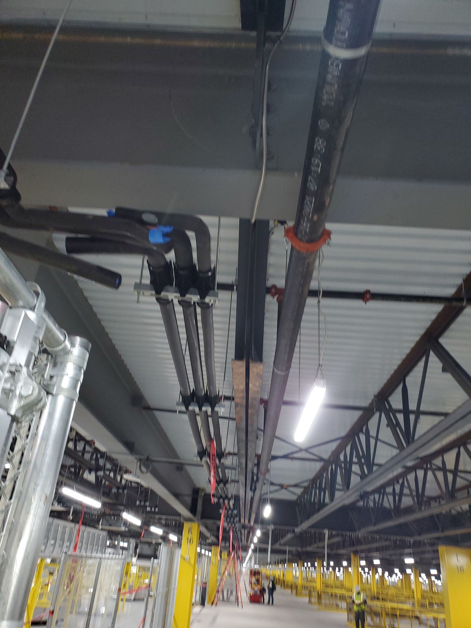 Overhead pipes and conduits in a warehouse with a high ceiling and a walkway below.