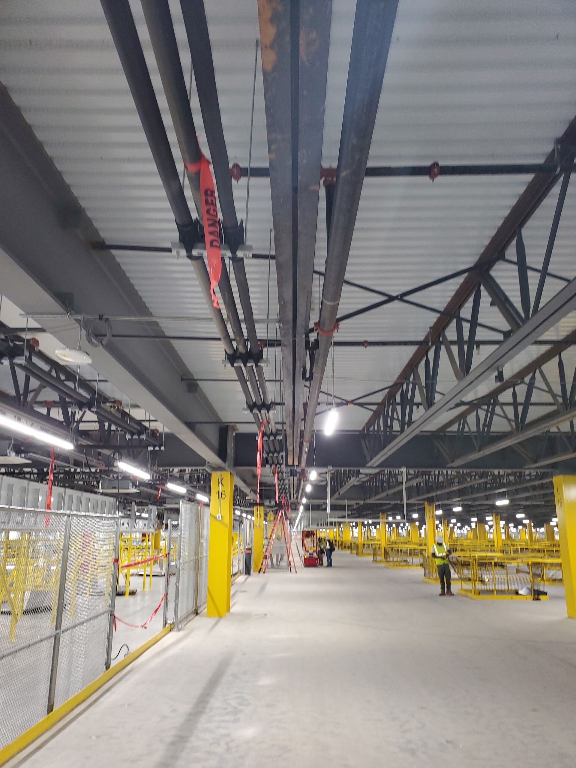 Warehouse interior with overhead conveyor system and yellow support structures. Workers in vests.