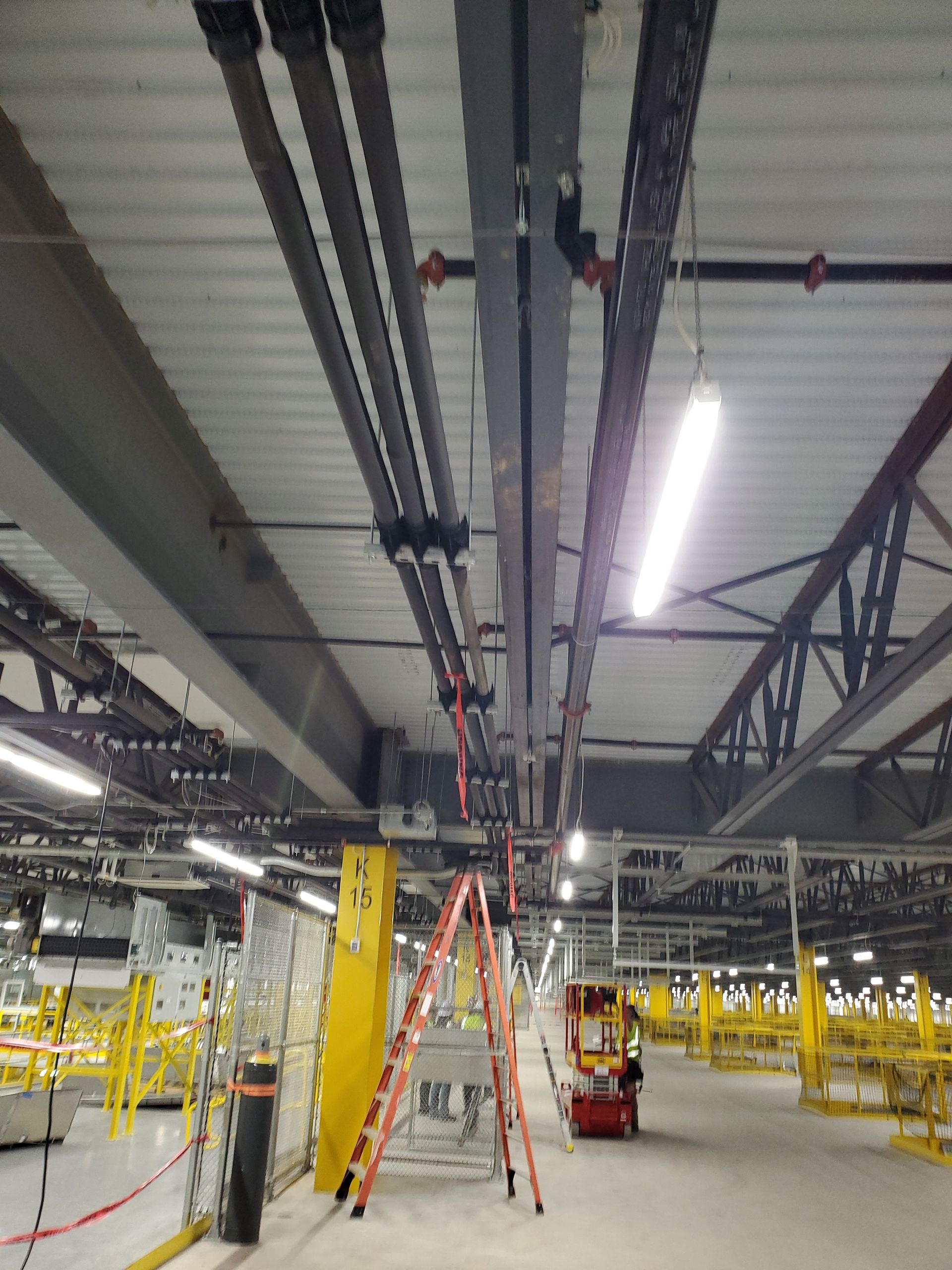 Warehouse ceiling with pipes, lights, and a ladder. Yellow structure and equipment below.