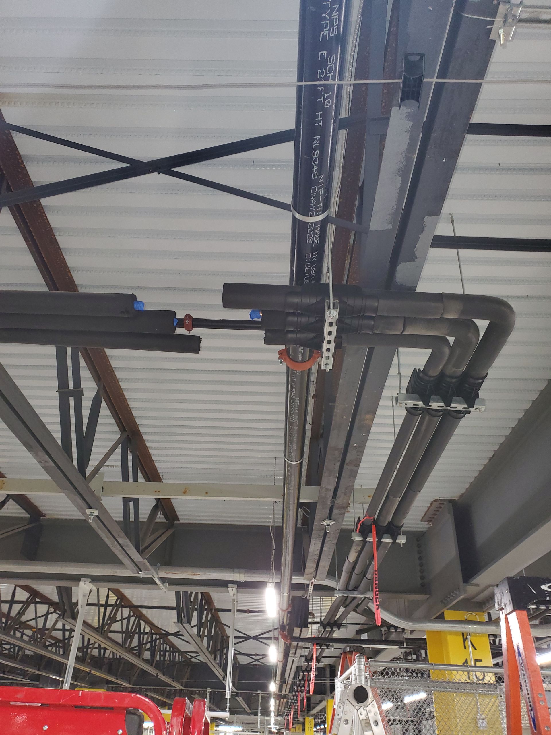 Overhead view of electrical conduit and wiring along a warehouse ceiling. White ceiling with metal beams and construction.