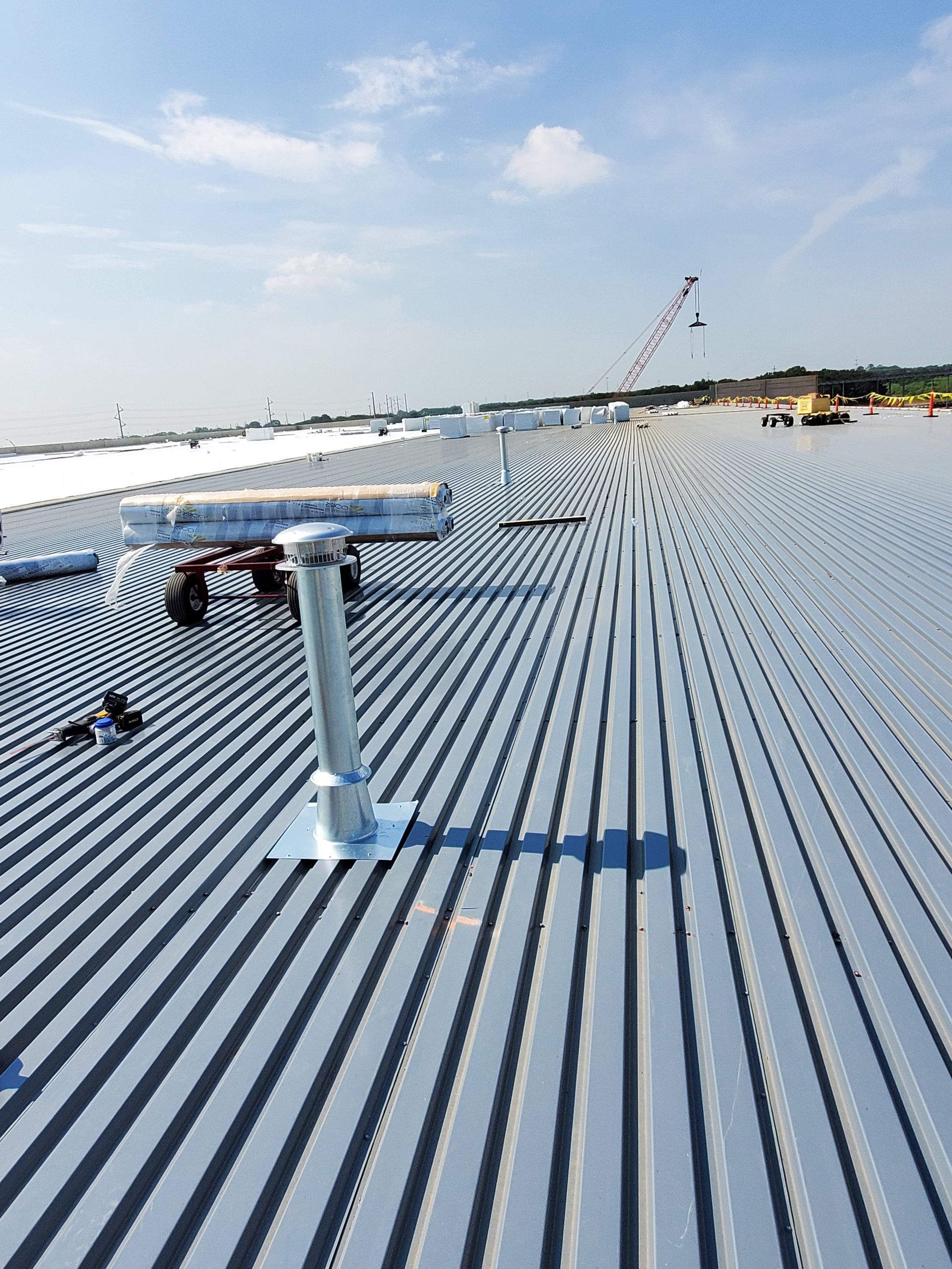 Metal roof with a vent and a distant crane under a blue sky.