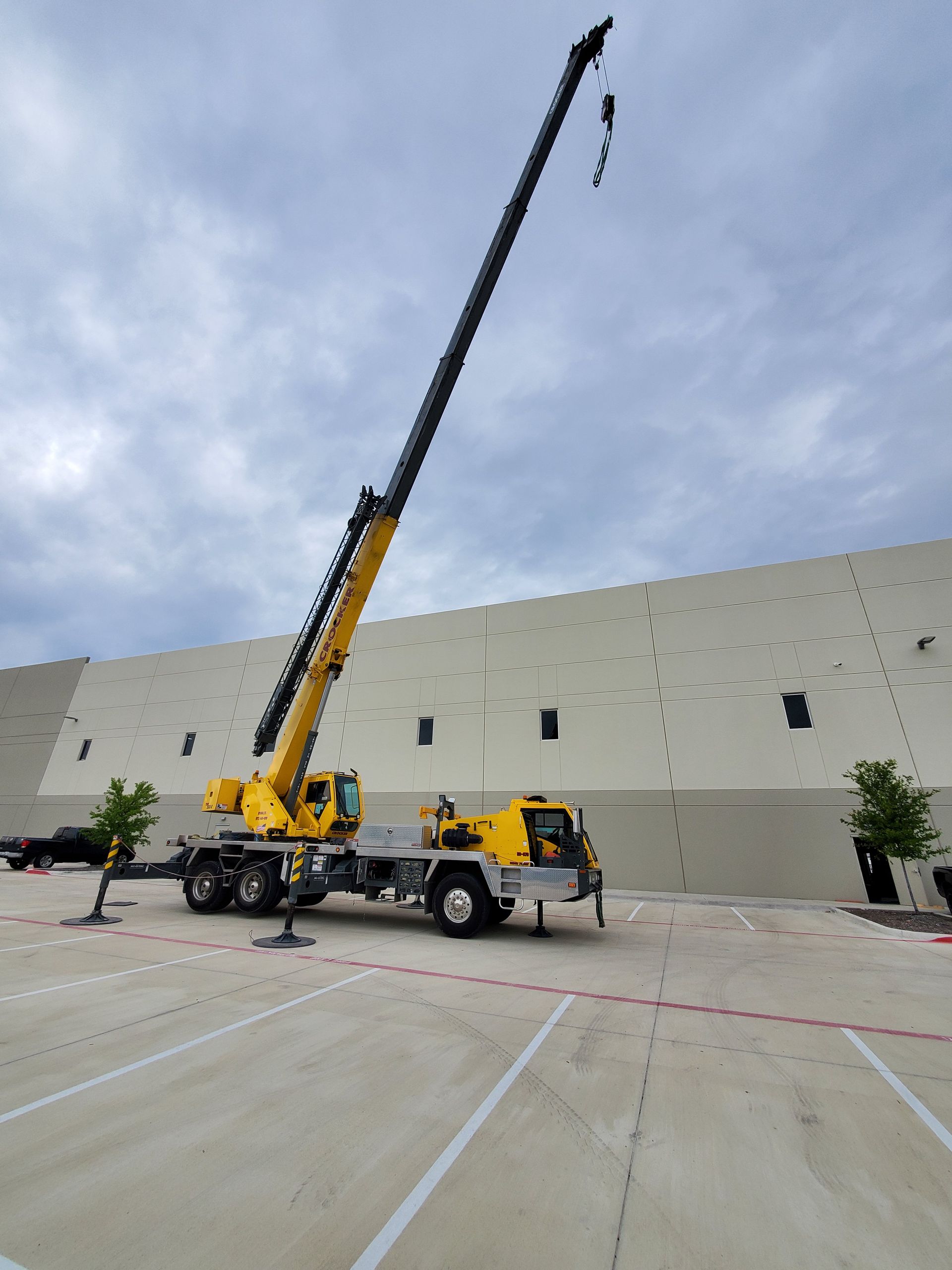 Yellow crane with an extended boom stands in a parking lot next to a large building under a cloudy sky.